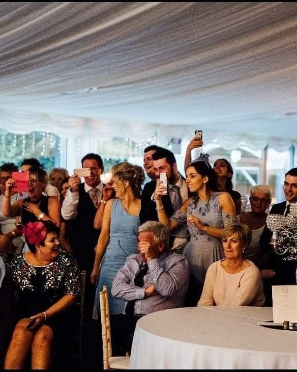 Guests at a wedding, some holding phones and capturing moments, seated under a decorated tent.
