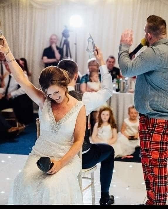 Bride and groom playing the shoe game at a wedding, surrounded by guests and a host with a microphone.