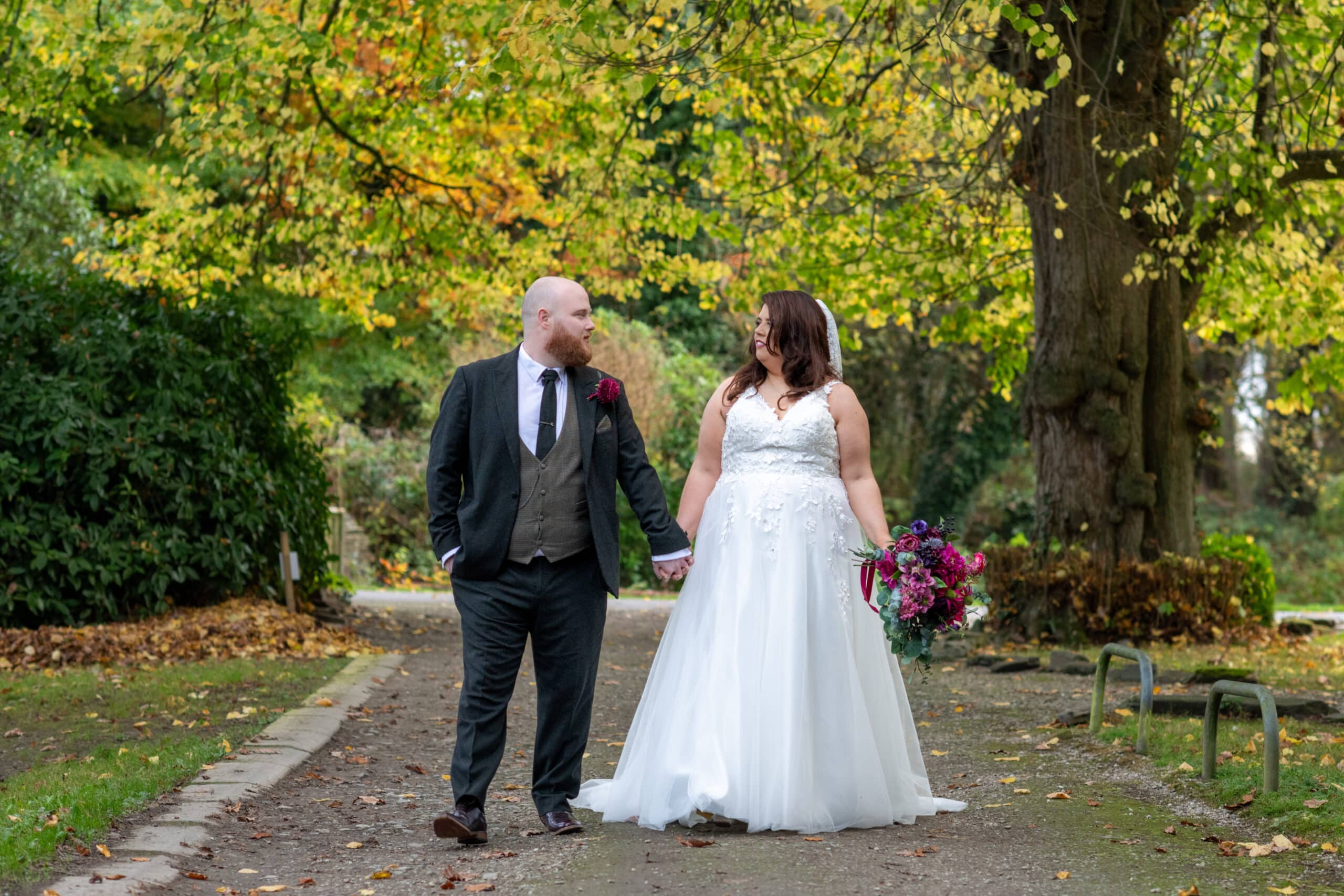 Bride and groom walking hand in hand on a tree-lined path, bride holding a bouquet.