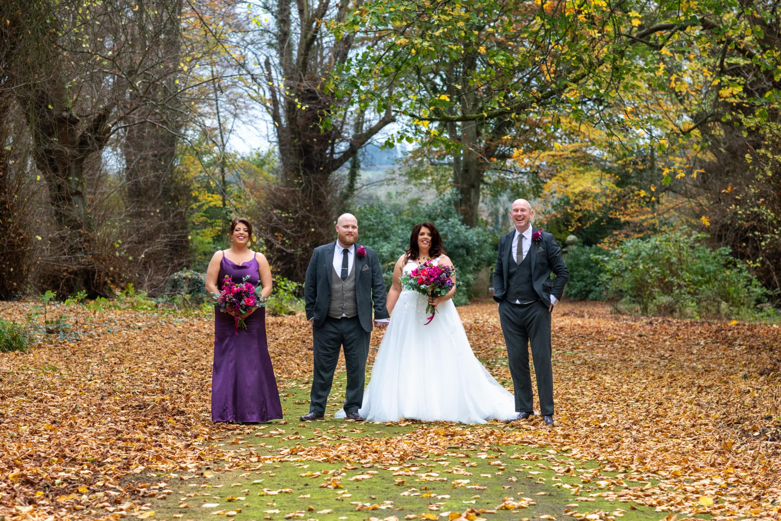Wedding party posing outdoors on a leaf-covered path with autumn trees in the background.