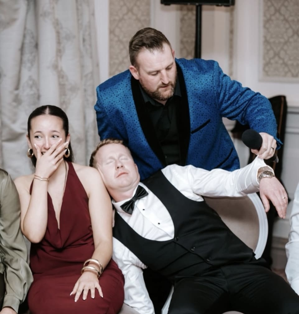 Man in blue suit holding microphone, woman laughing, another man in formal attire leaning asleep.