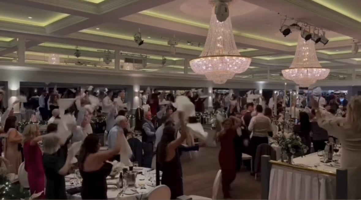 Guests waving napkins at a formal event under large chandeliers.