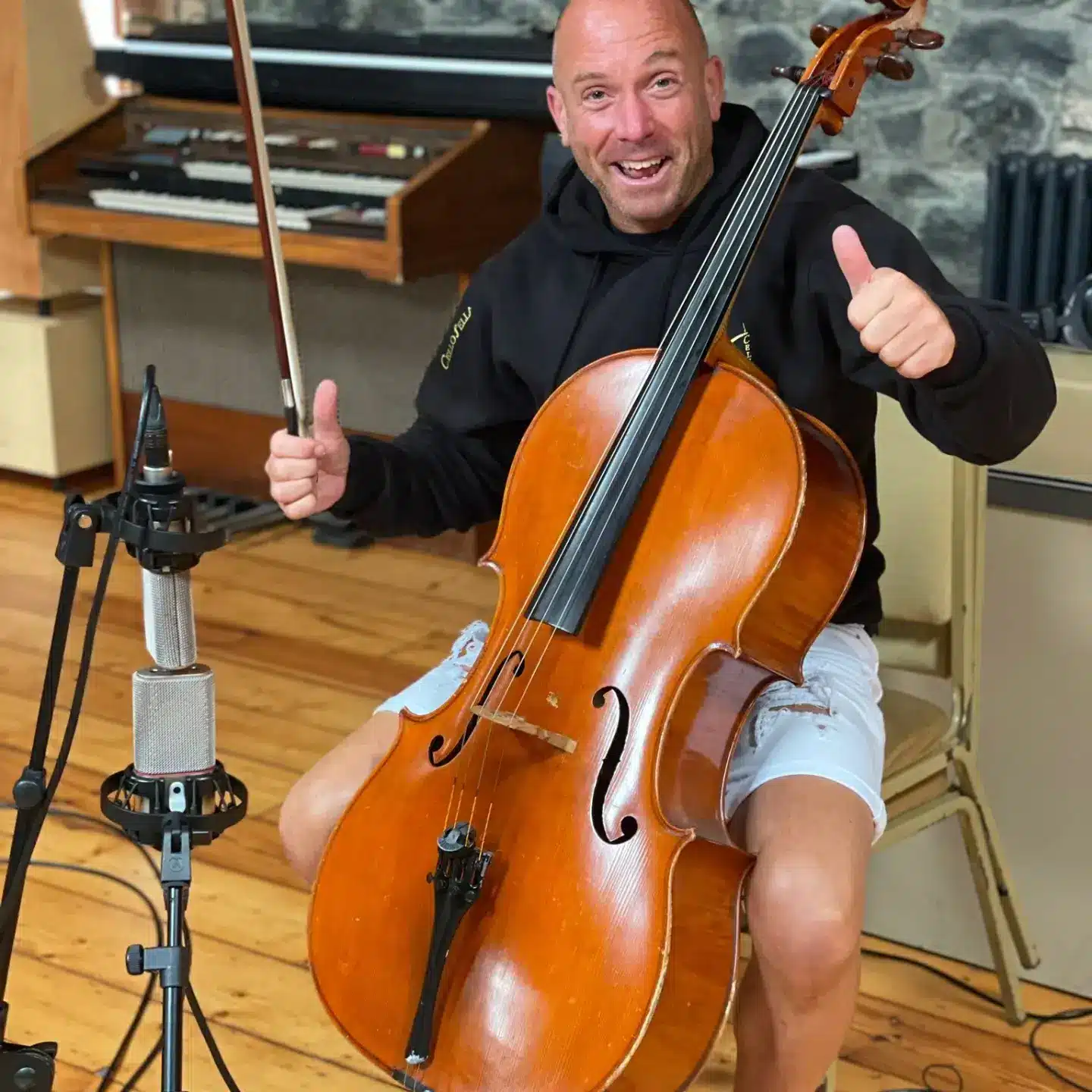 Man smiling while holding a cello and giving a thumbs-up in a music studio.