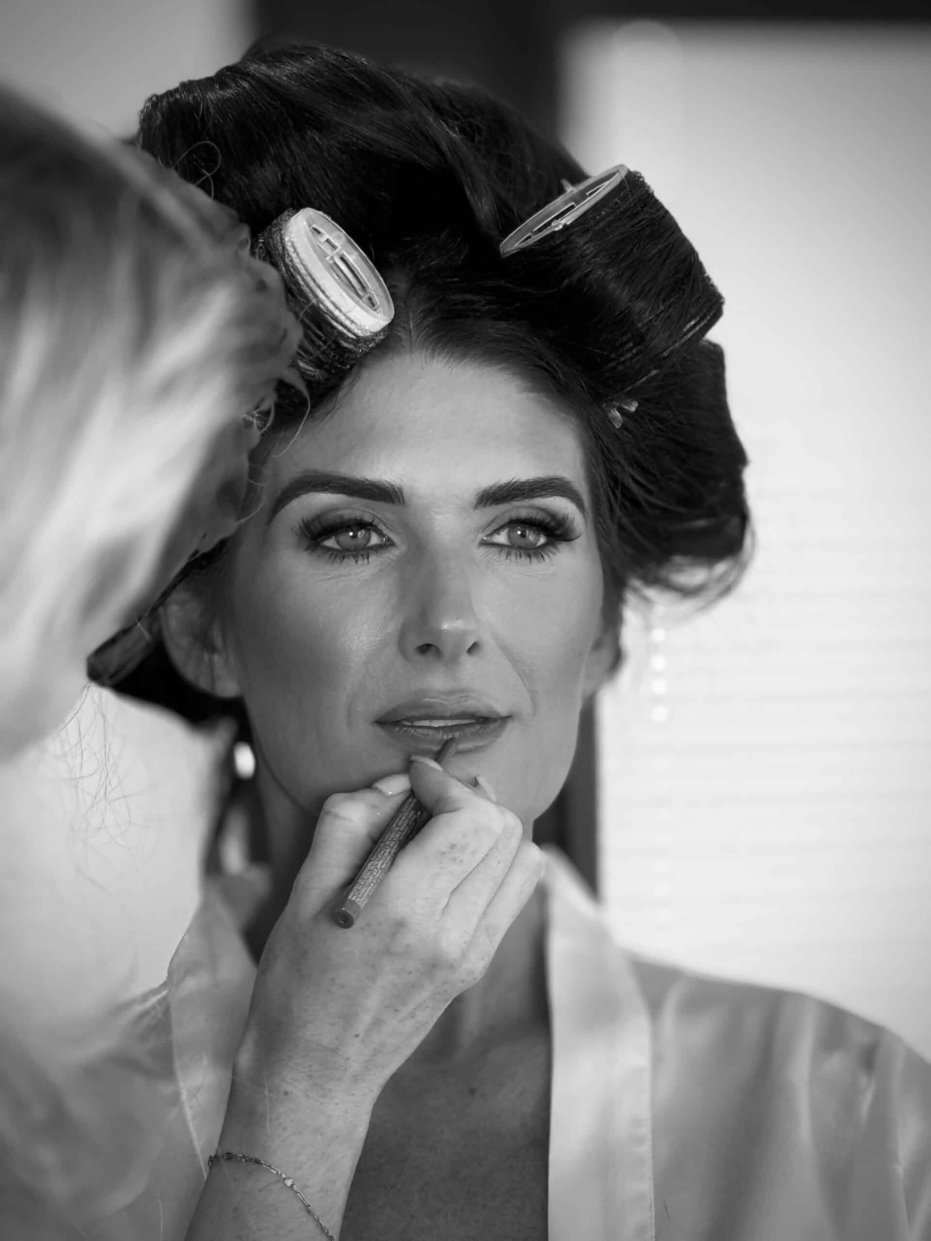 Woman having makeup applied, wearing hair rollers, black and white photo.