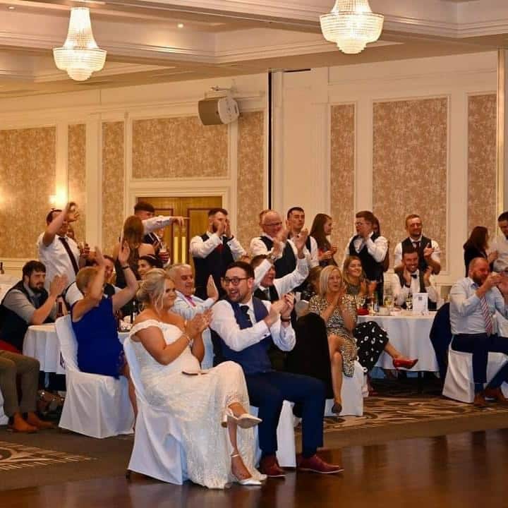 Guests in formal attire clapping and celebrating at an indoor event, possibly a wedding reception.