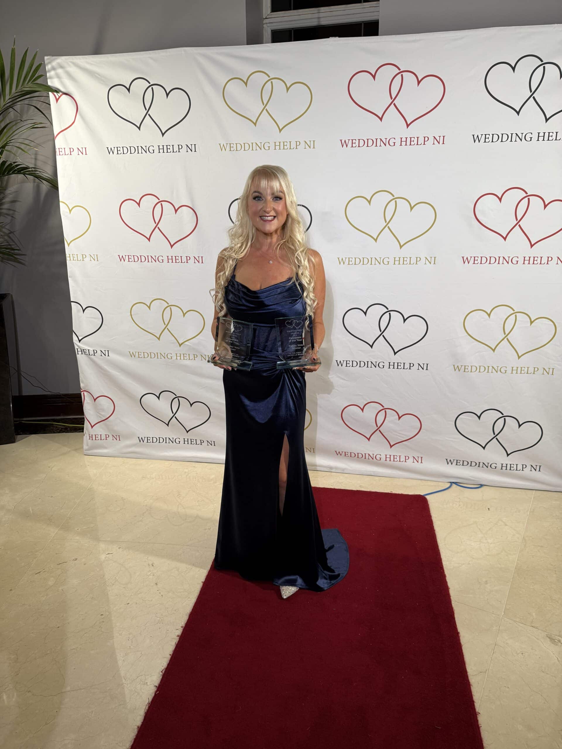 Woman in blue dress holding awards, standing on red carpet in front of "Wedding Help NI" backdrop.