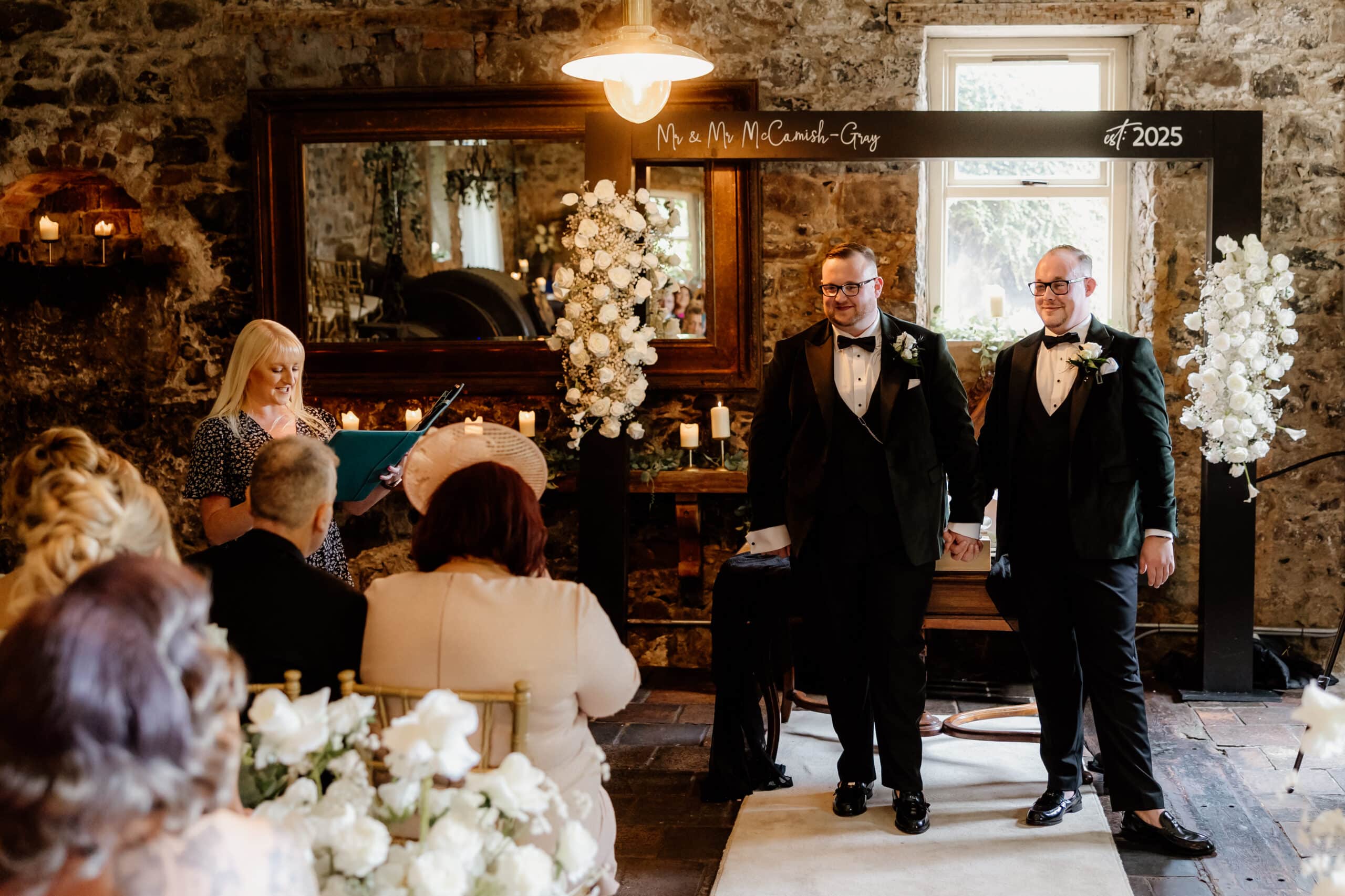 Two grooms holding hands at their wedding ceremony, with a celebrant and guests seated in a rustic venue.