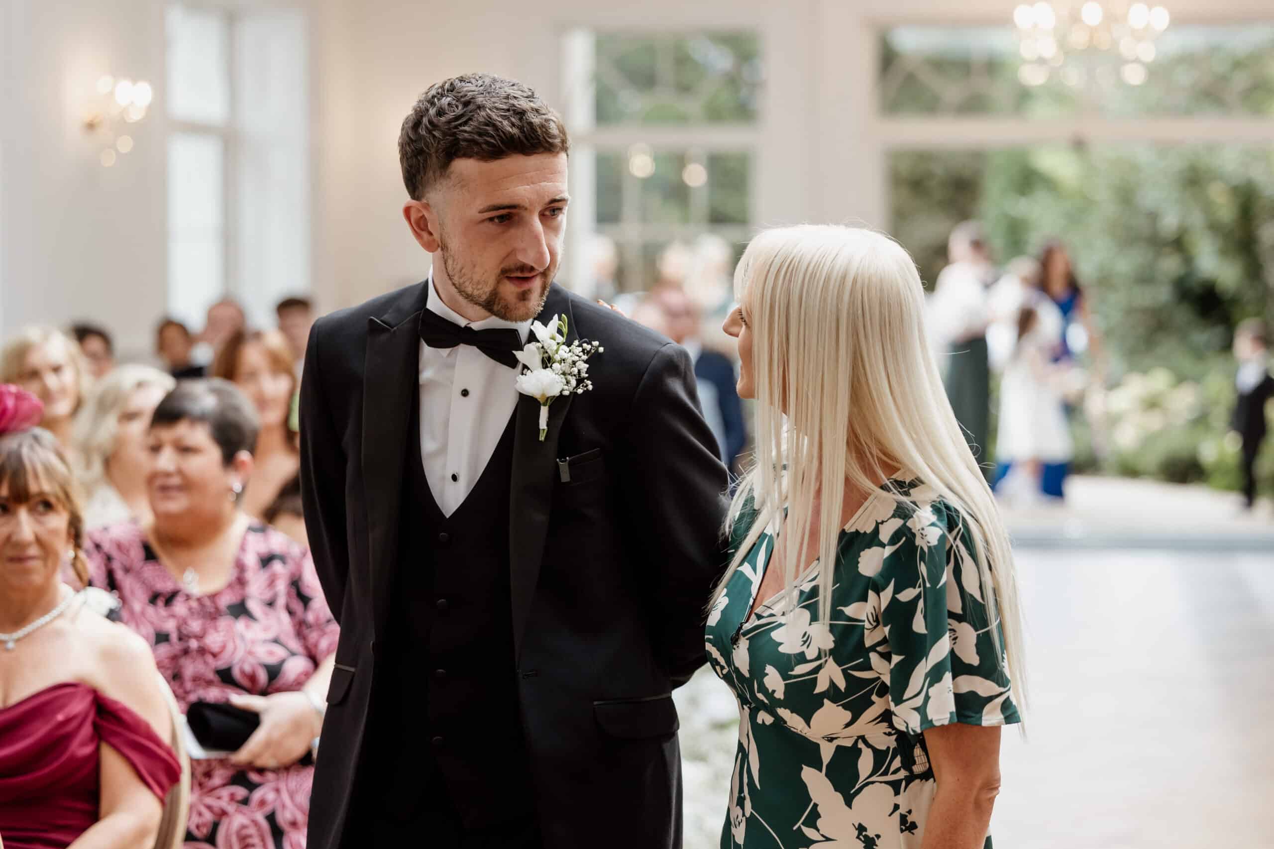A man in a tuxedo and woman in a floral dress at an indoor event, with an audience in the background.