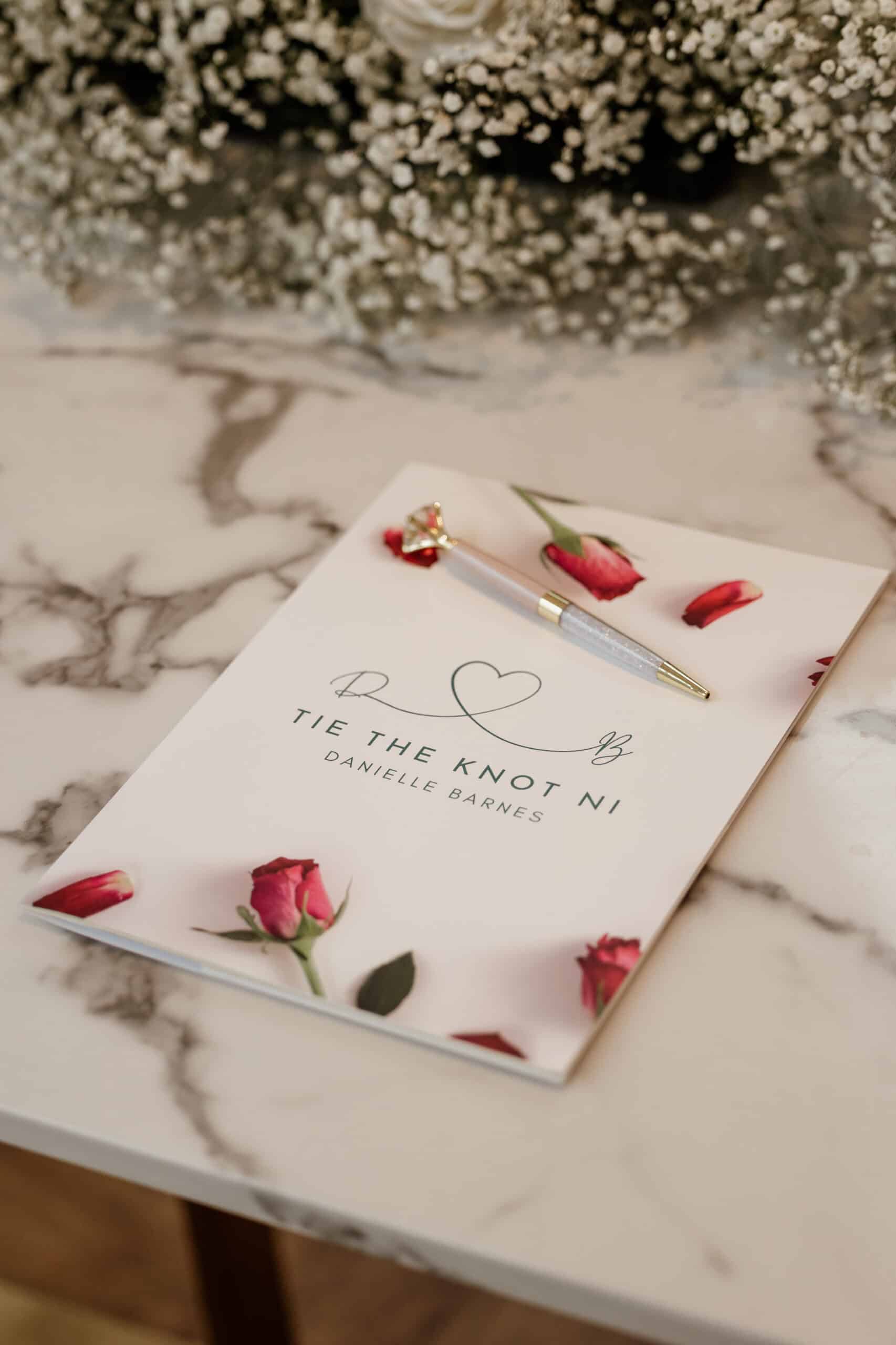 Wedding planning booklet on a marble table with rose petals and a decorative pen.