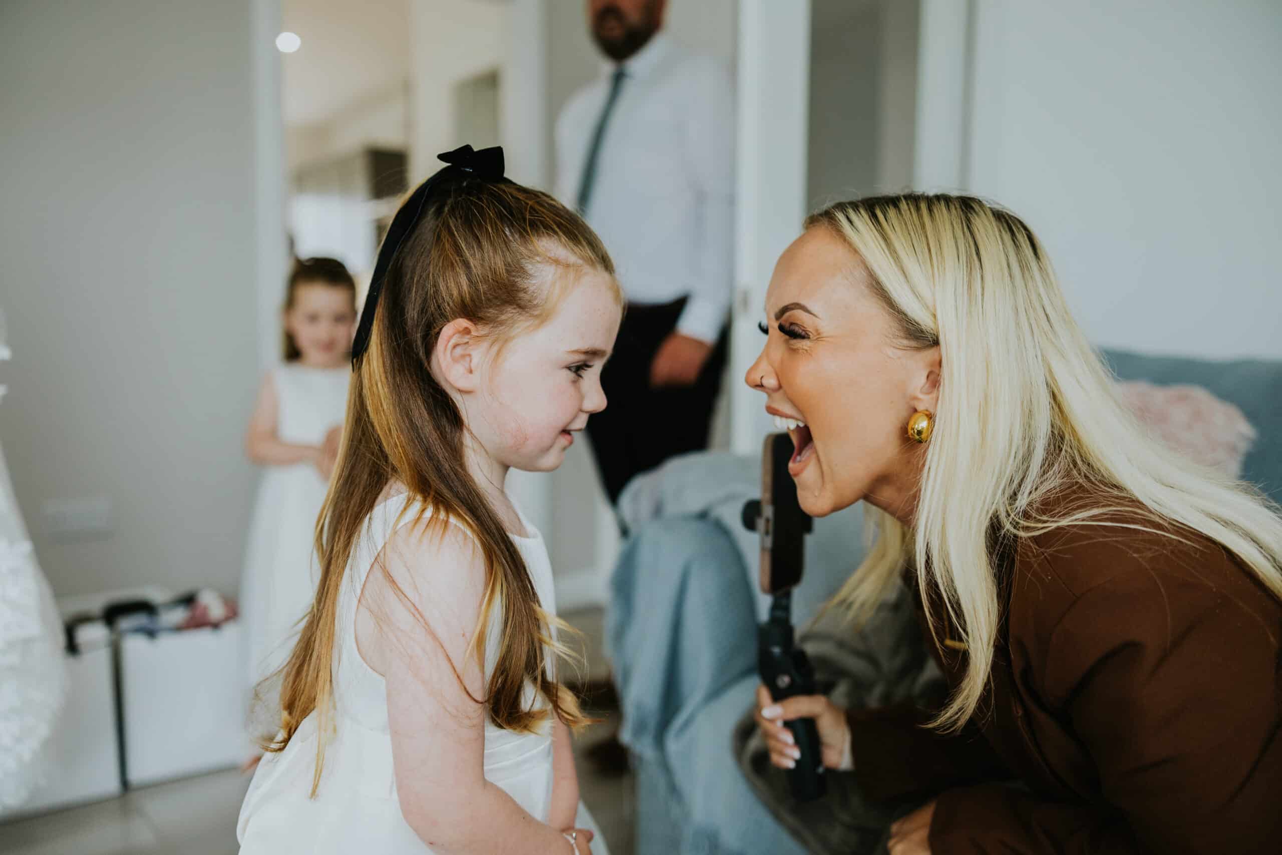 Woman and young girl smiling at each other indoors, both dressed in formal attire.