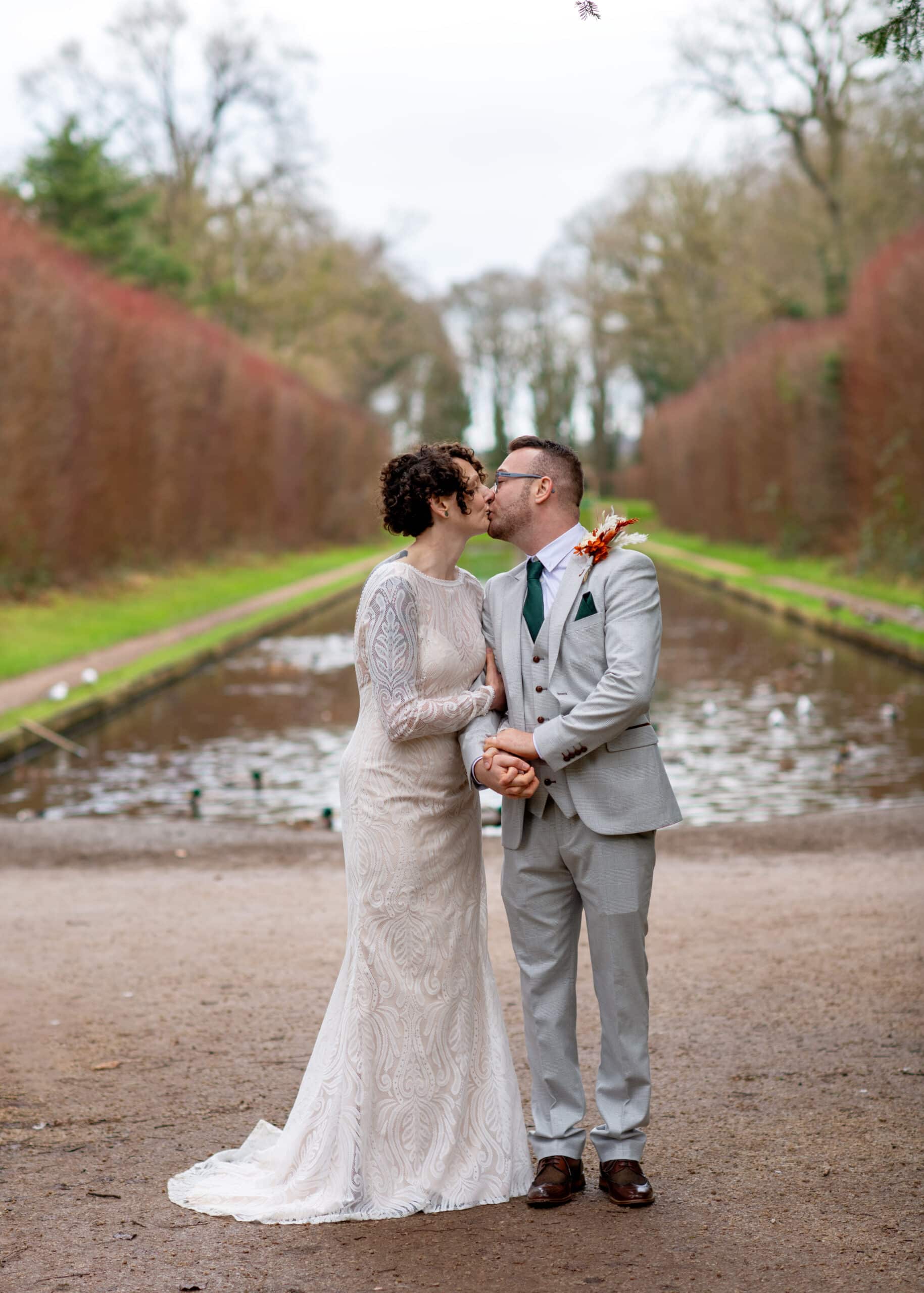 Couple kissing in wedding attire by a scenic pond with trees in the background.
