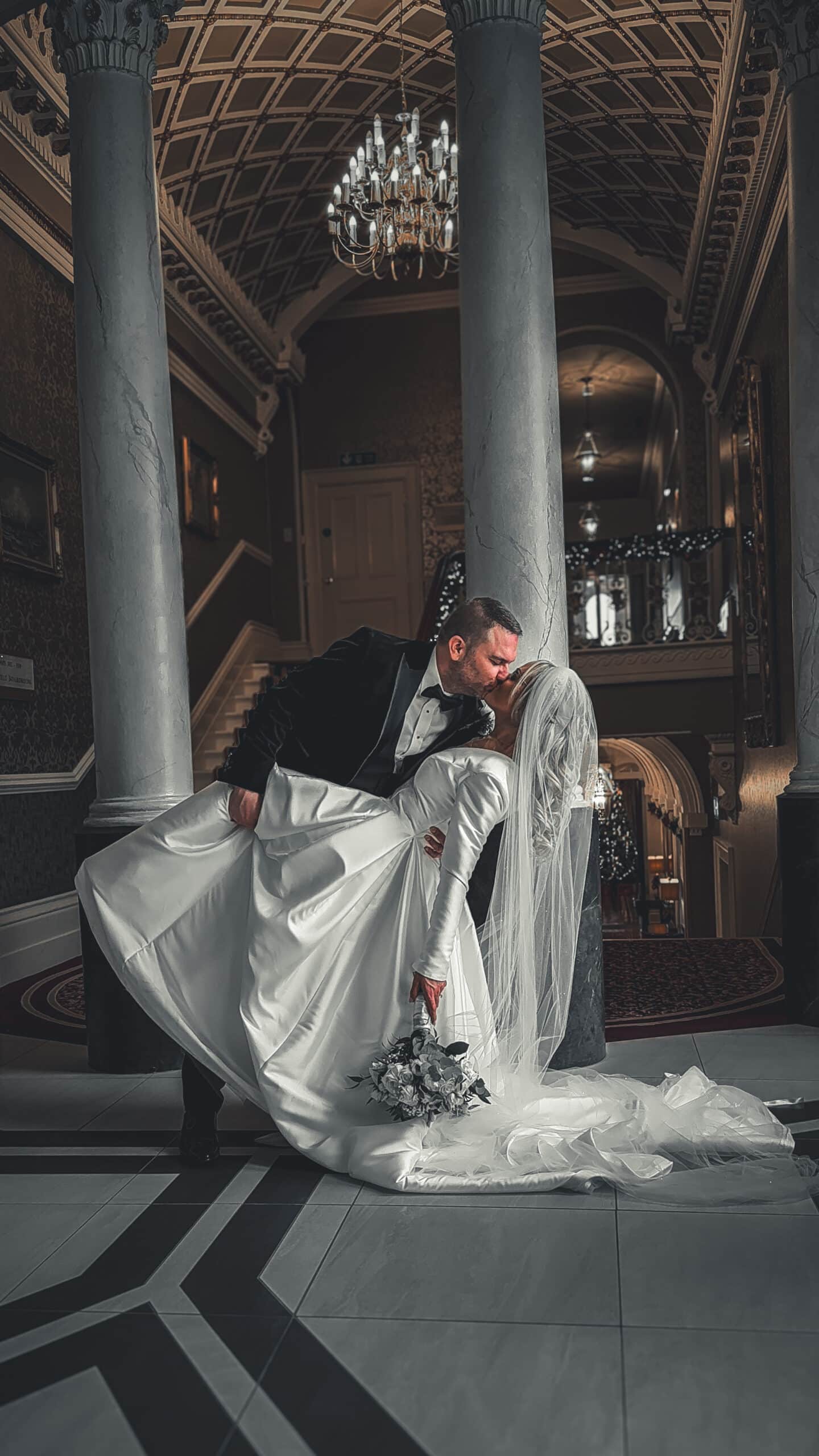 Bride and groom share a kiss in a grand, elegant hallway with a chandelier.