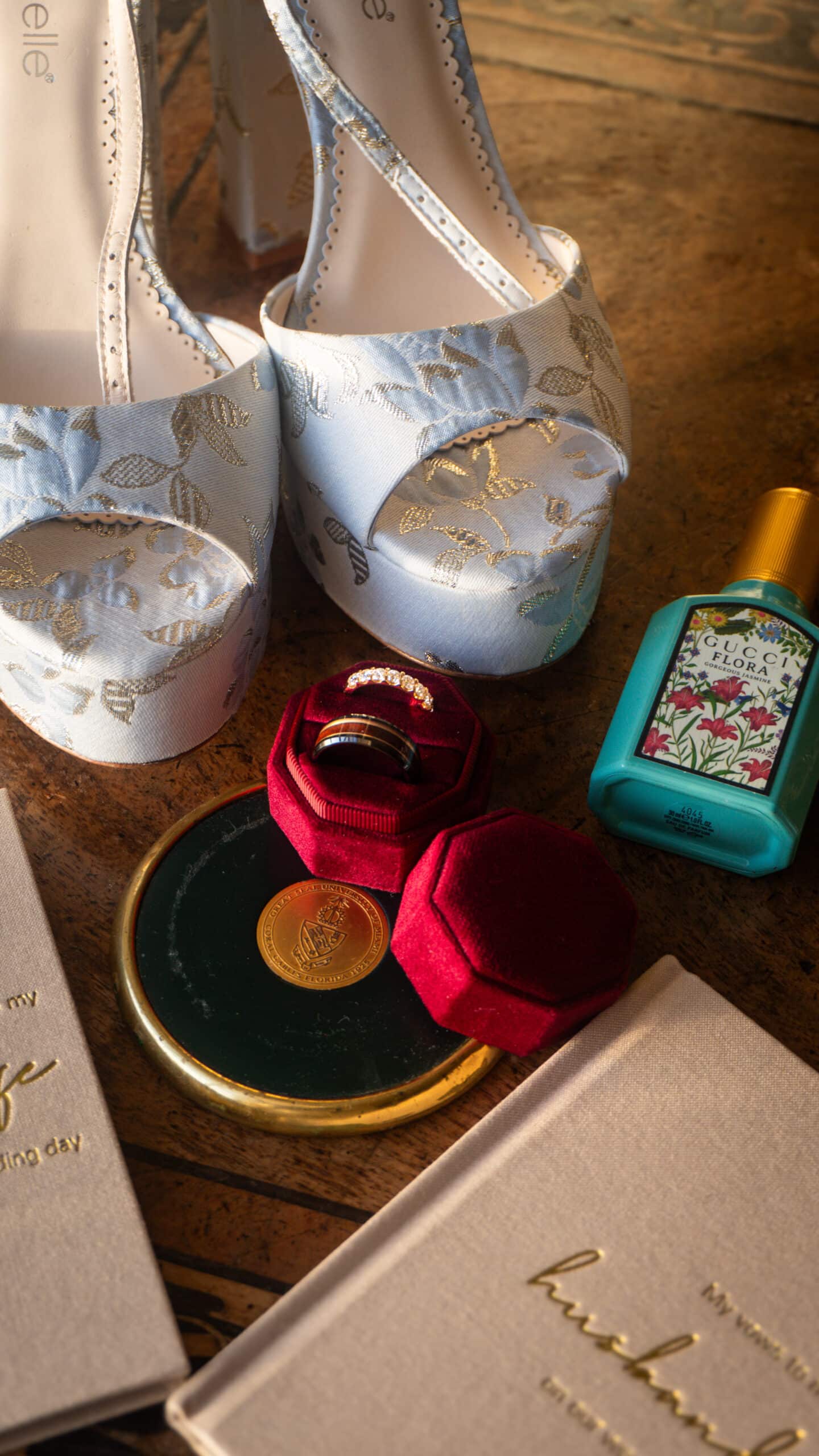 Perfume bottle, decorative shoes, velvet ring box, and vow books on a wooden surface.