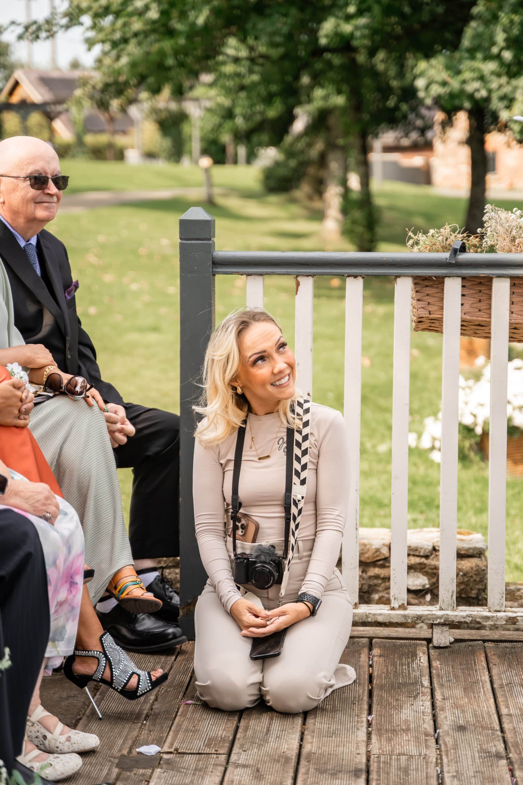 Woman kneeling with a camera, smiling outdoors near a wooden fence, surrounded by people in formal attire.
