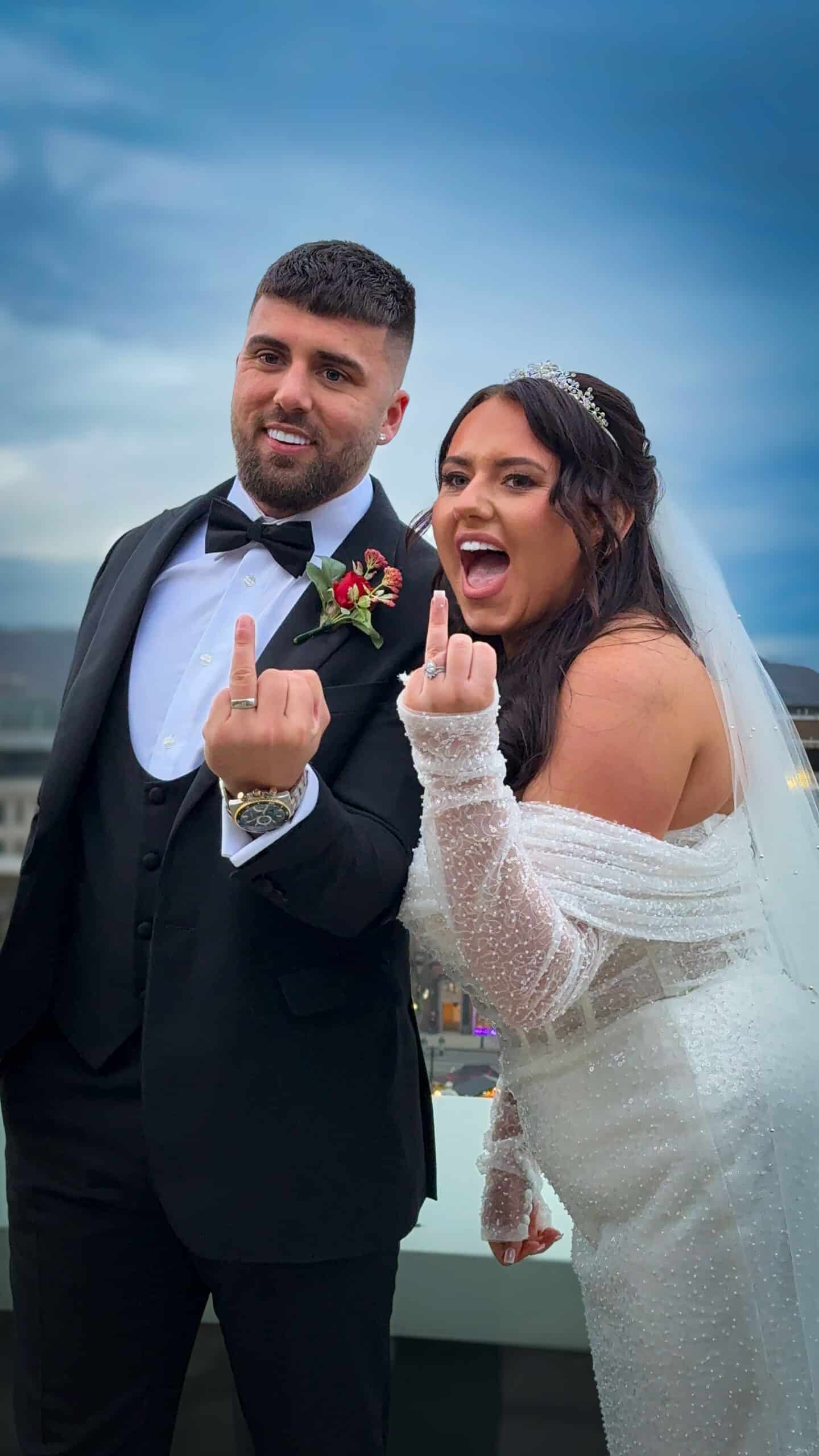 Bride and groom in wedding attire, both smiling and holding up middle fingers against an outdoor backdrop.