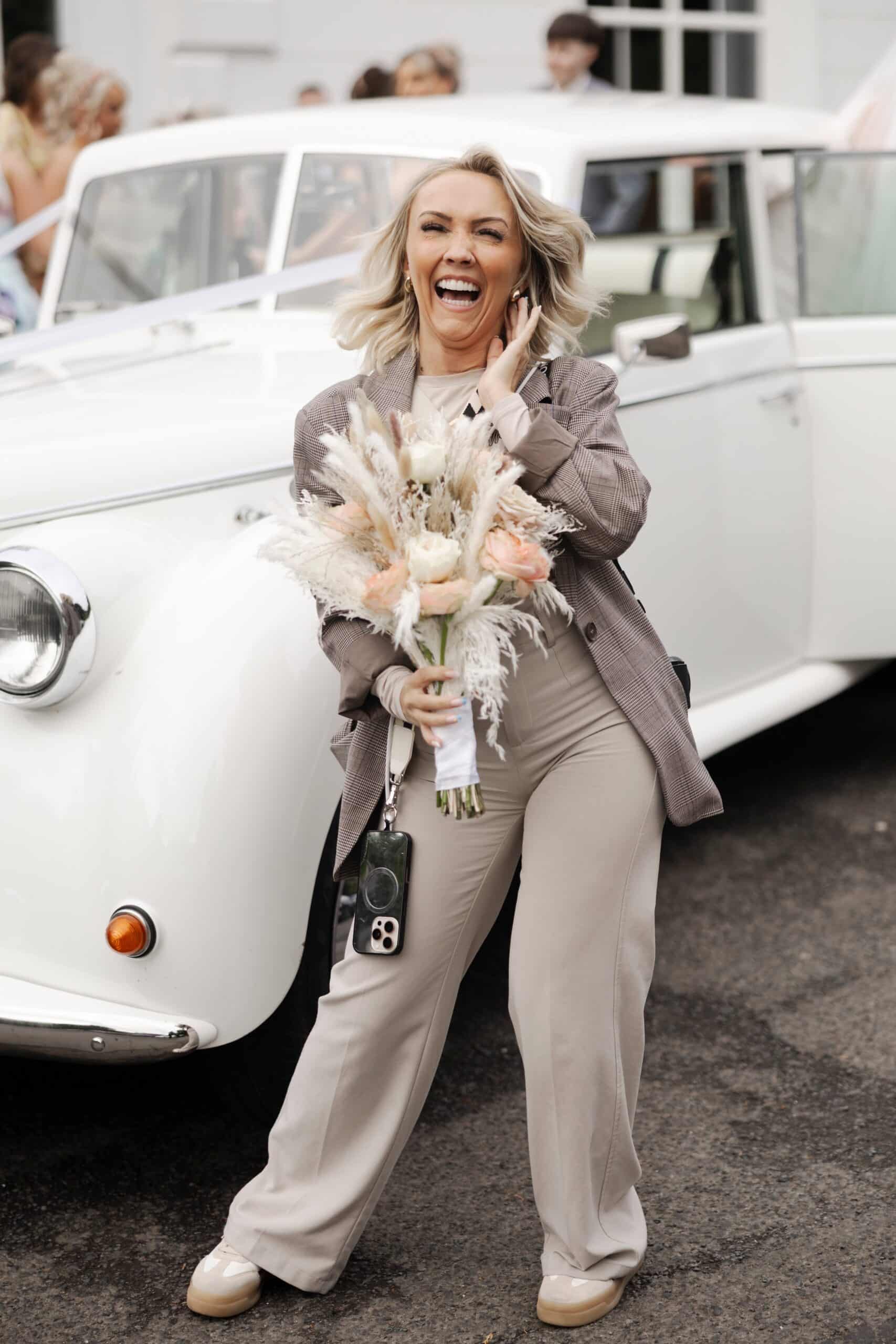 Woman smiling and holding bouquet in front of a classic white car, wearing neutral-toned outfit.