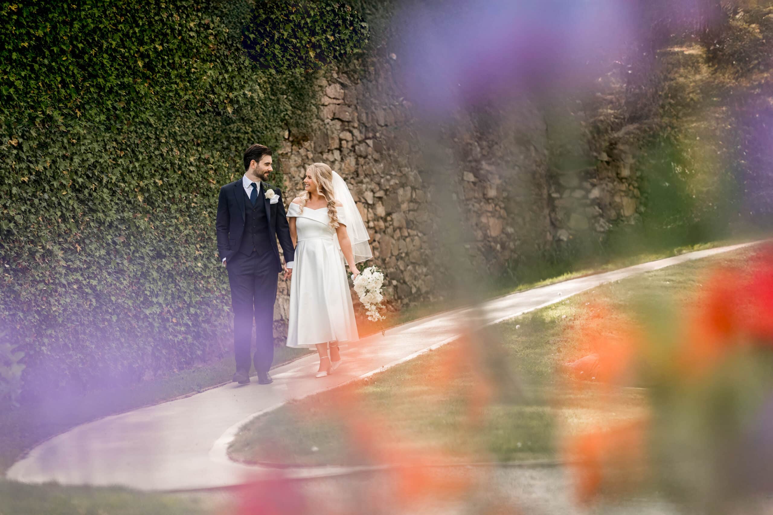 Bride and groom walking on a pathway surrounded by flowers and greenery, holding hands.