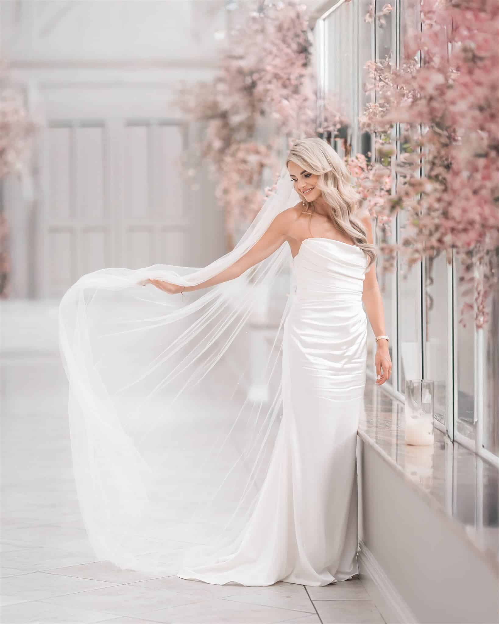 Bride in elegant white gown with veil, standing in a bright hallway adorned with pink flowers.