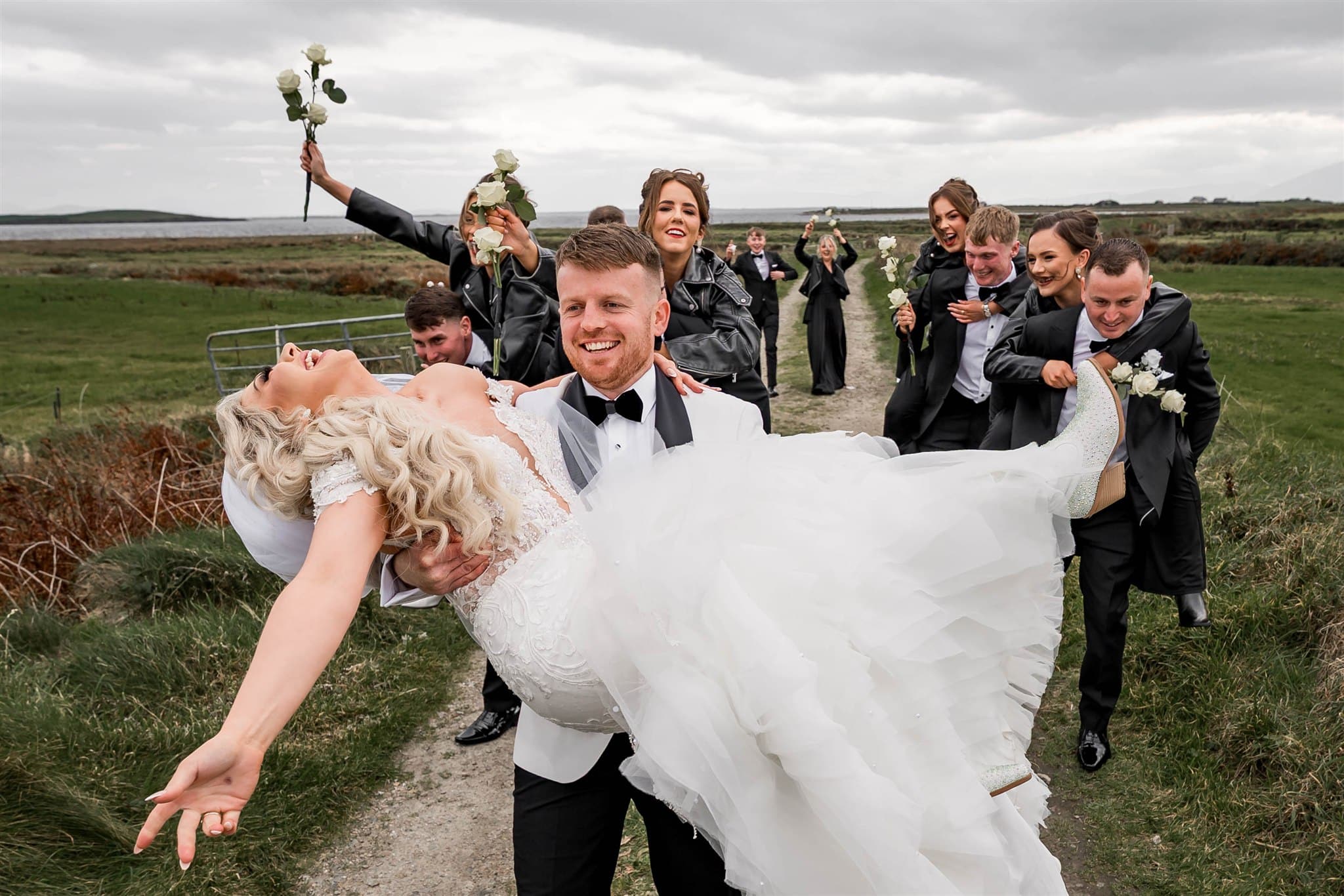 Bride and groom joyfully pose with bridal party outdoors, groom holding bride as groomsmen and bridesmaids cheer with roses.