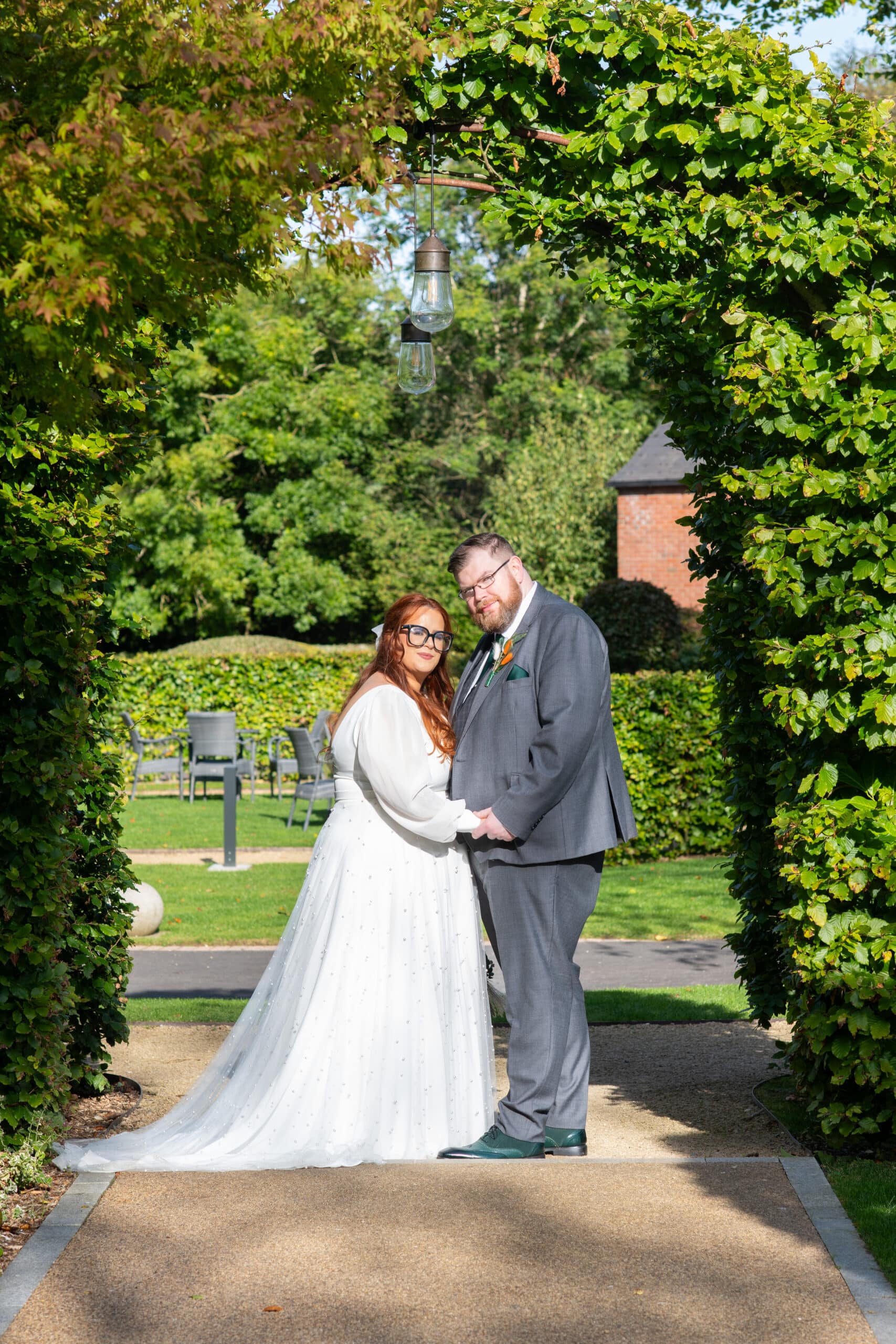 Bride and groom standing under a lush green archway in a garden setting.