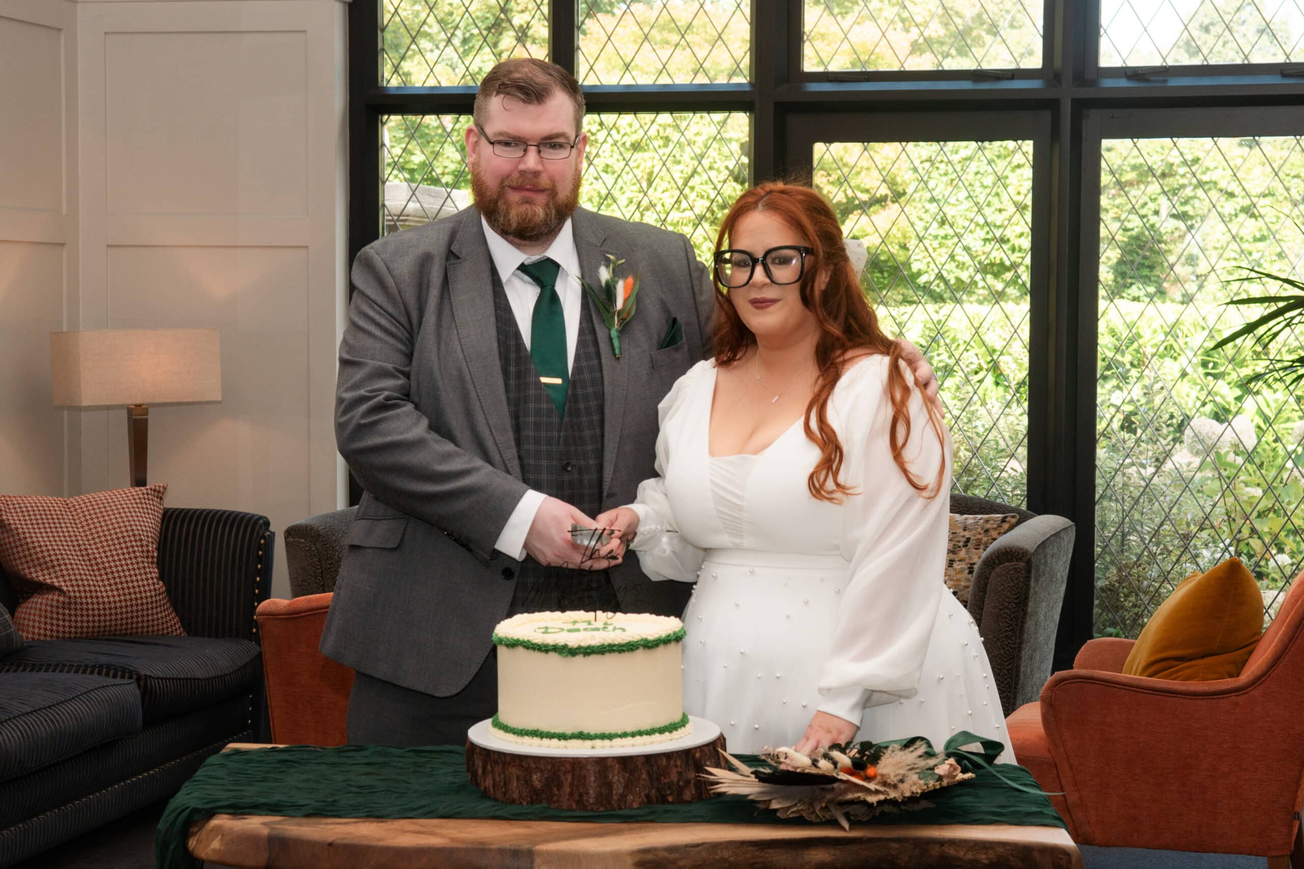Couple cutting a wedding cake in a room with large windows and decorative furniture.
