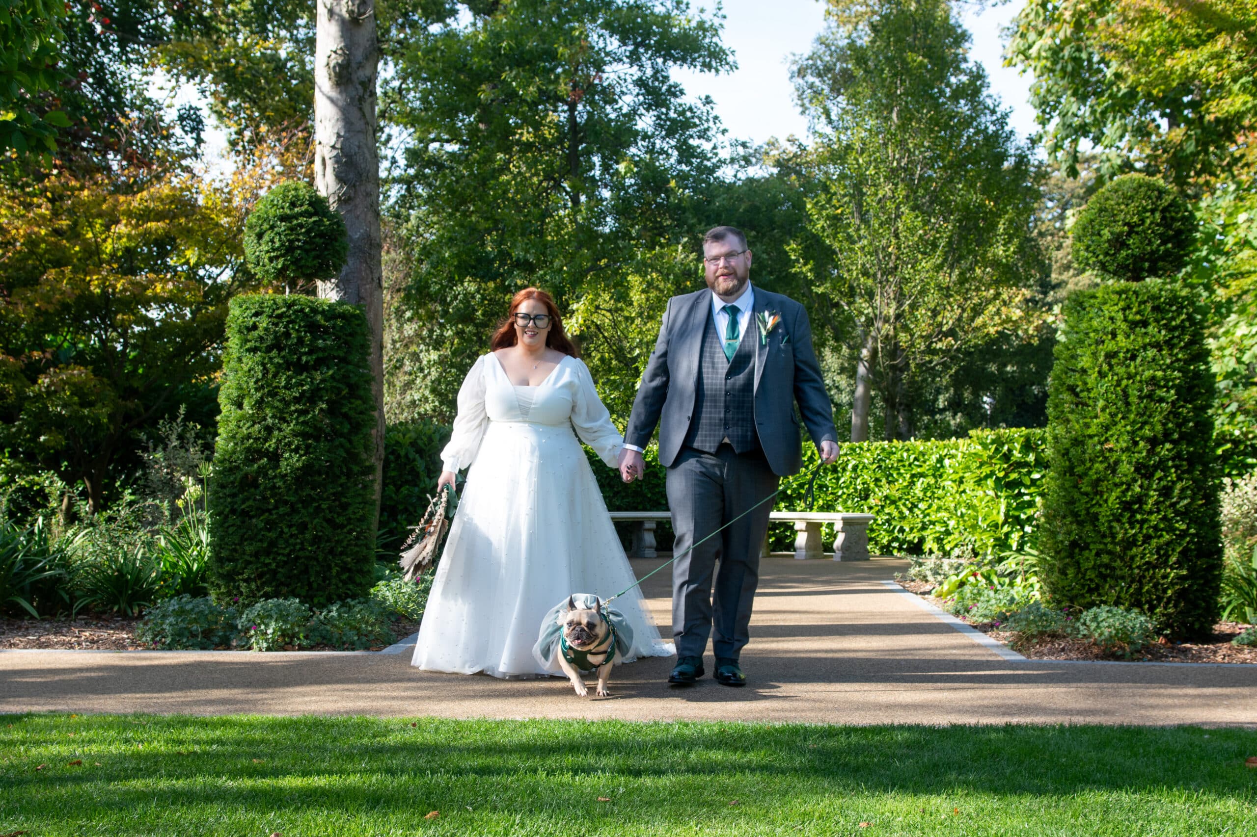 A couple in wedding attire walks their dog down a garden path on a sunny day.