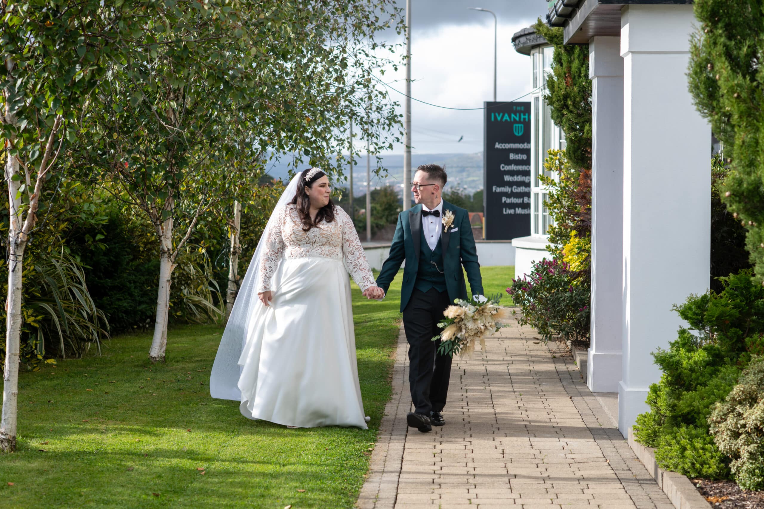 Bride and groom walking hand in hand outside a venue with greenery and a sign in the background.