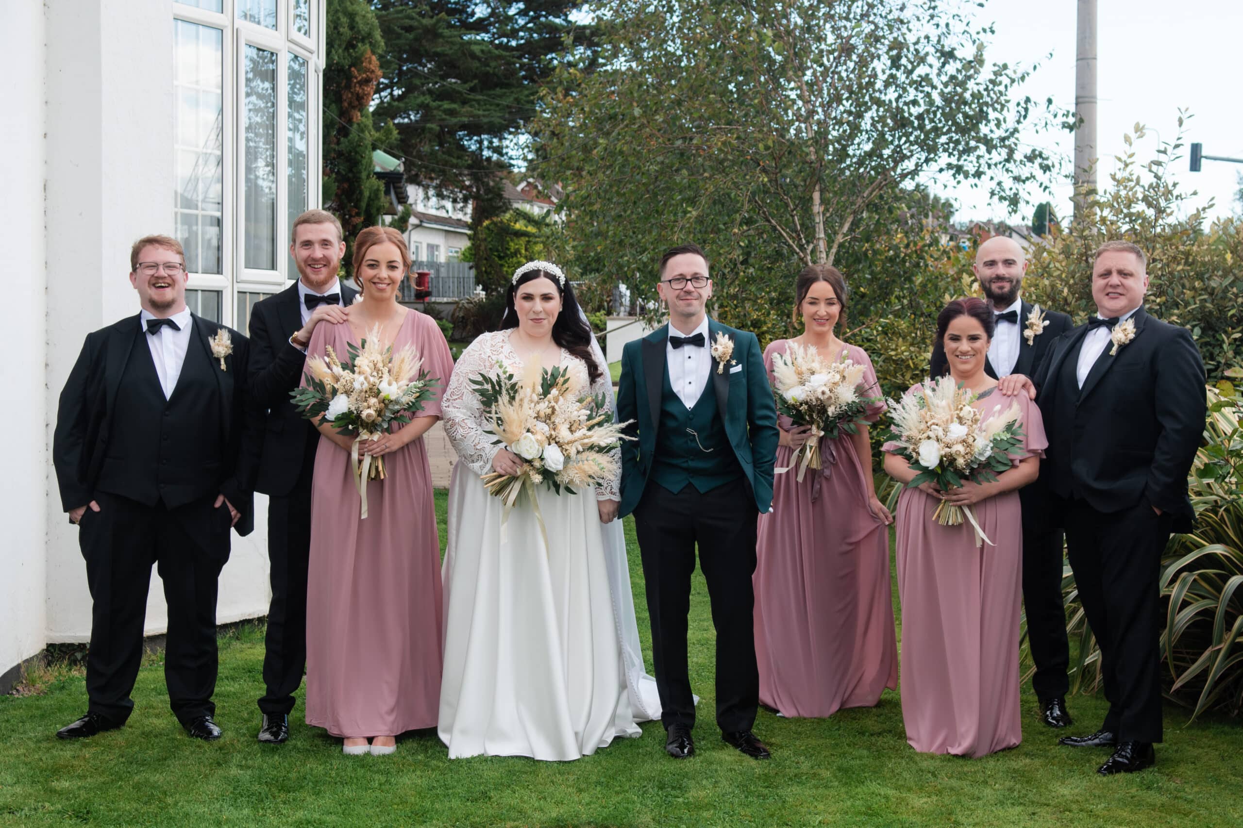 Bride, groom, and wedding party pose outdoors in elegant attire with floral bouquets.