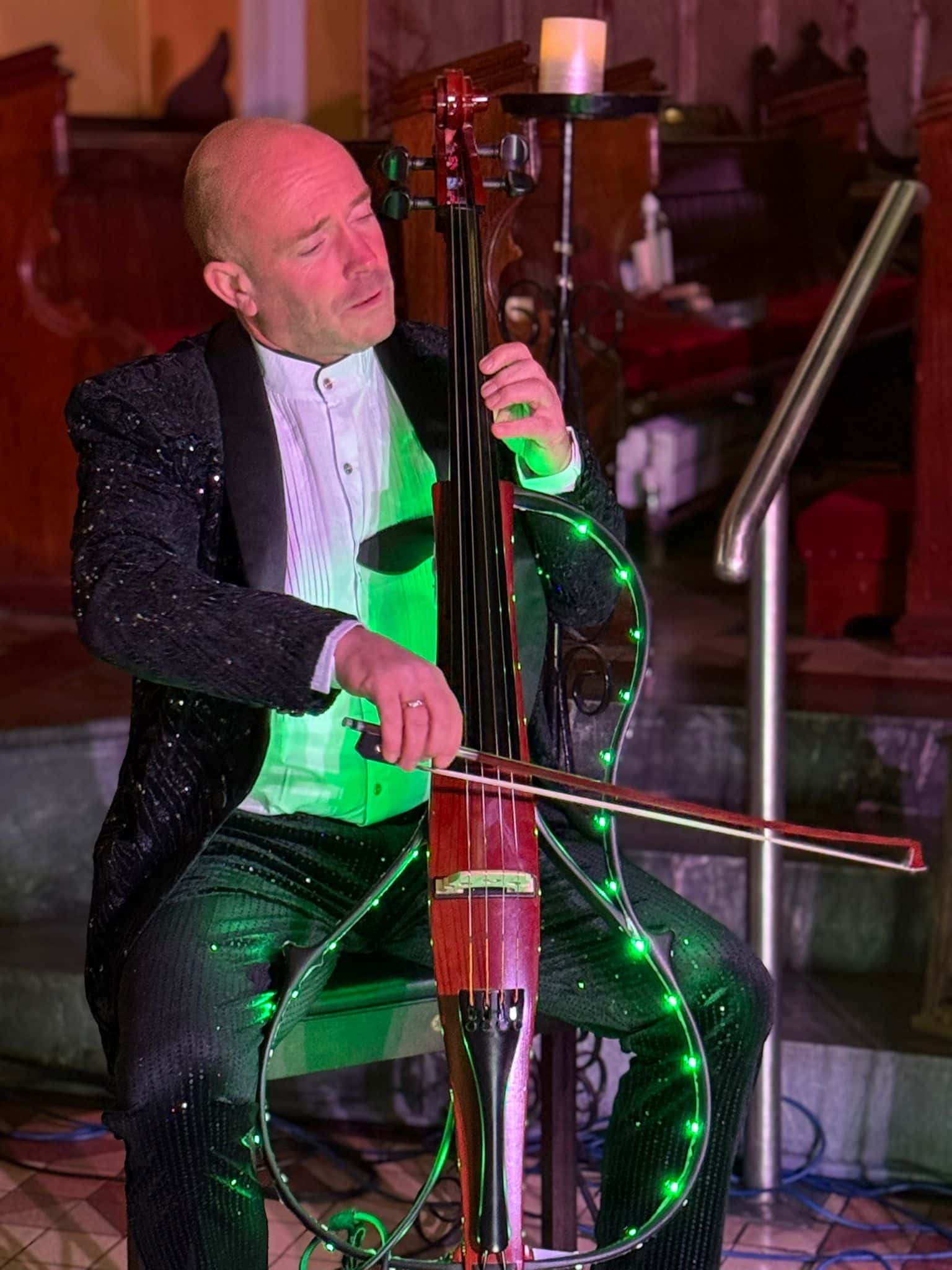 Musician playing a cello adorned with green lights, wearing a black sequined suit in a dimly lit room.
