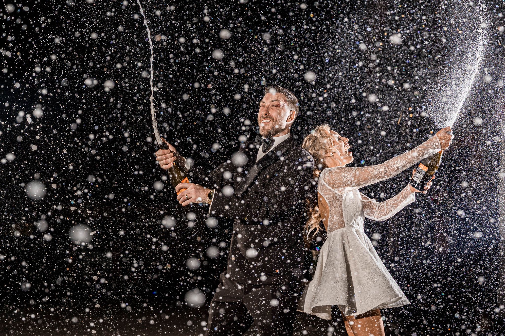 Couple celebrating with champagne, laughing amidst flying champagne droplets.