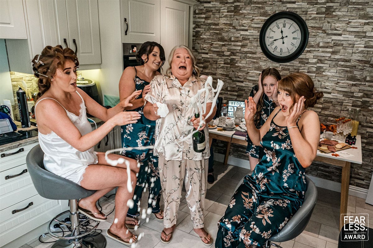 Group of women in a kitchen celebrating, with champagne foaming over.