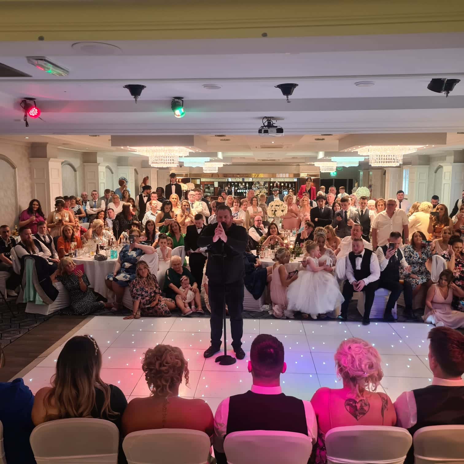 A crowded wedding reception with a man speaking into a microphone; guests are seated and standing around elegantly decorated tables.