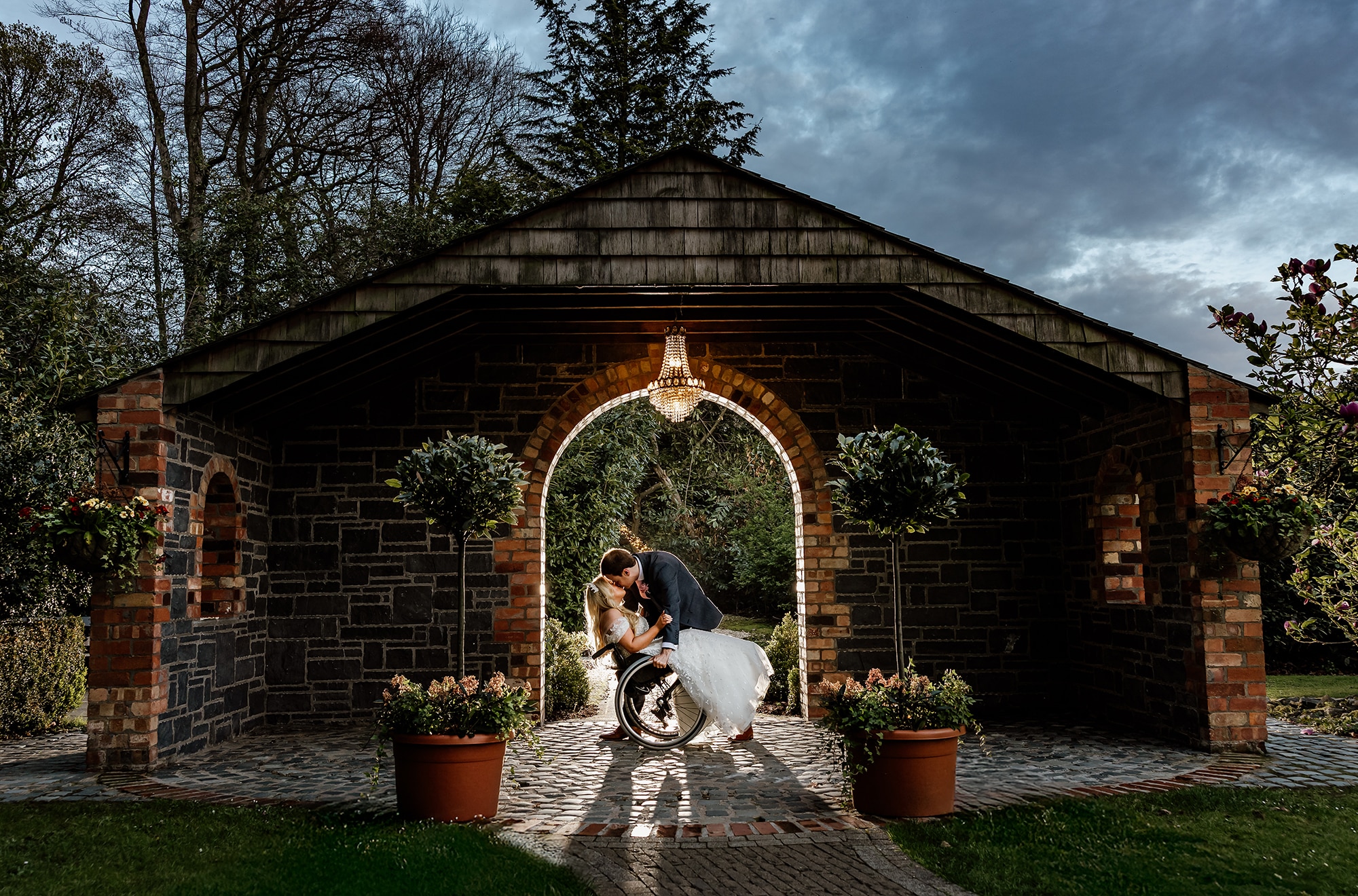 Bride and groom sharing a kiss under a rustic archway, chandelier above, surrounded by greenery and potted plants.
