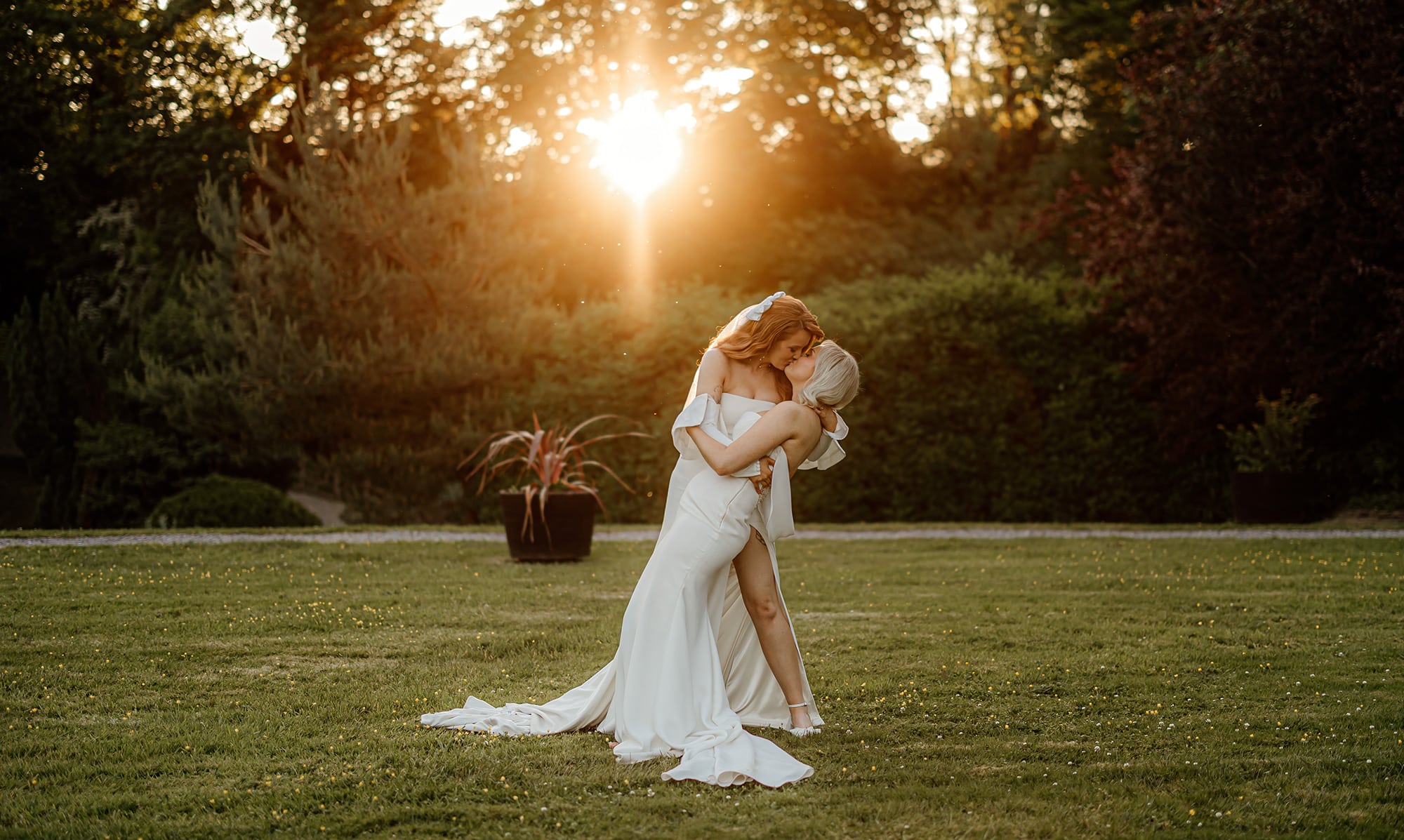 Two people in wedding attire kissing passionately in a sunlit garden.