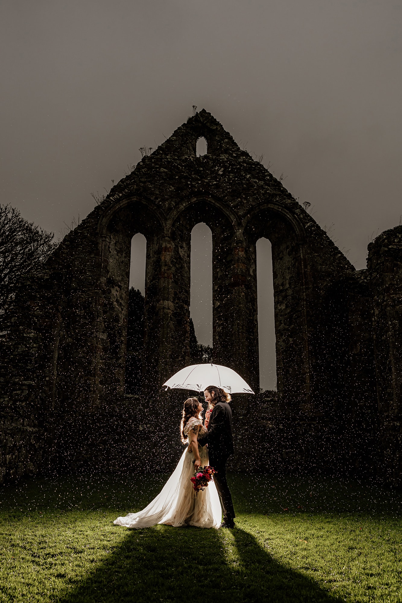 Bride and groom under umbrella in front of ruins, illuminated in rain.