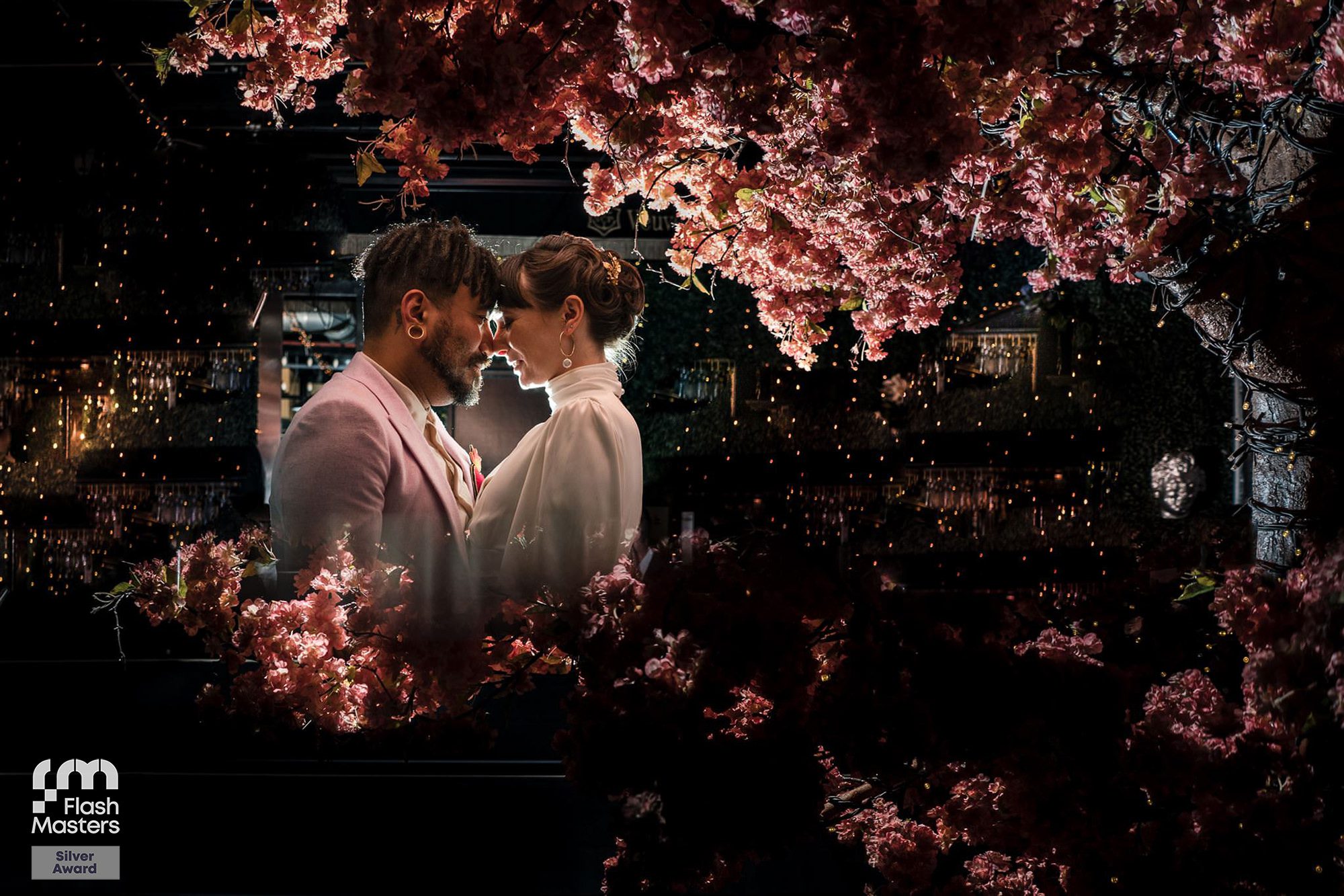 Couple embracing under cherry blossoms with decorative lights, Flash Masters Silver Award logo in corner.
