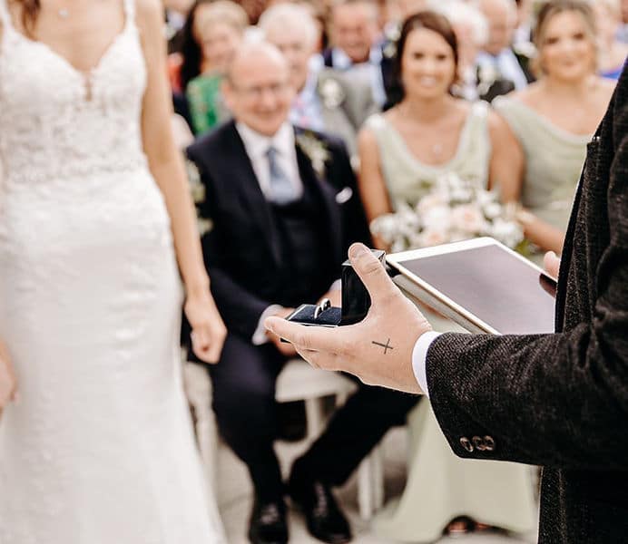 Close-up of a wedding ceremony with a groom holding a ring box and vow book, bride visible, guests seated in the background.