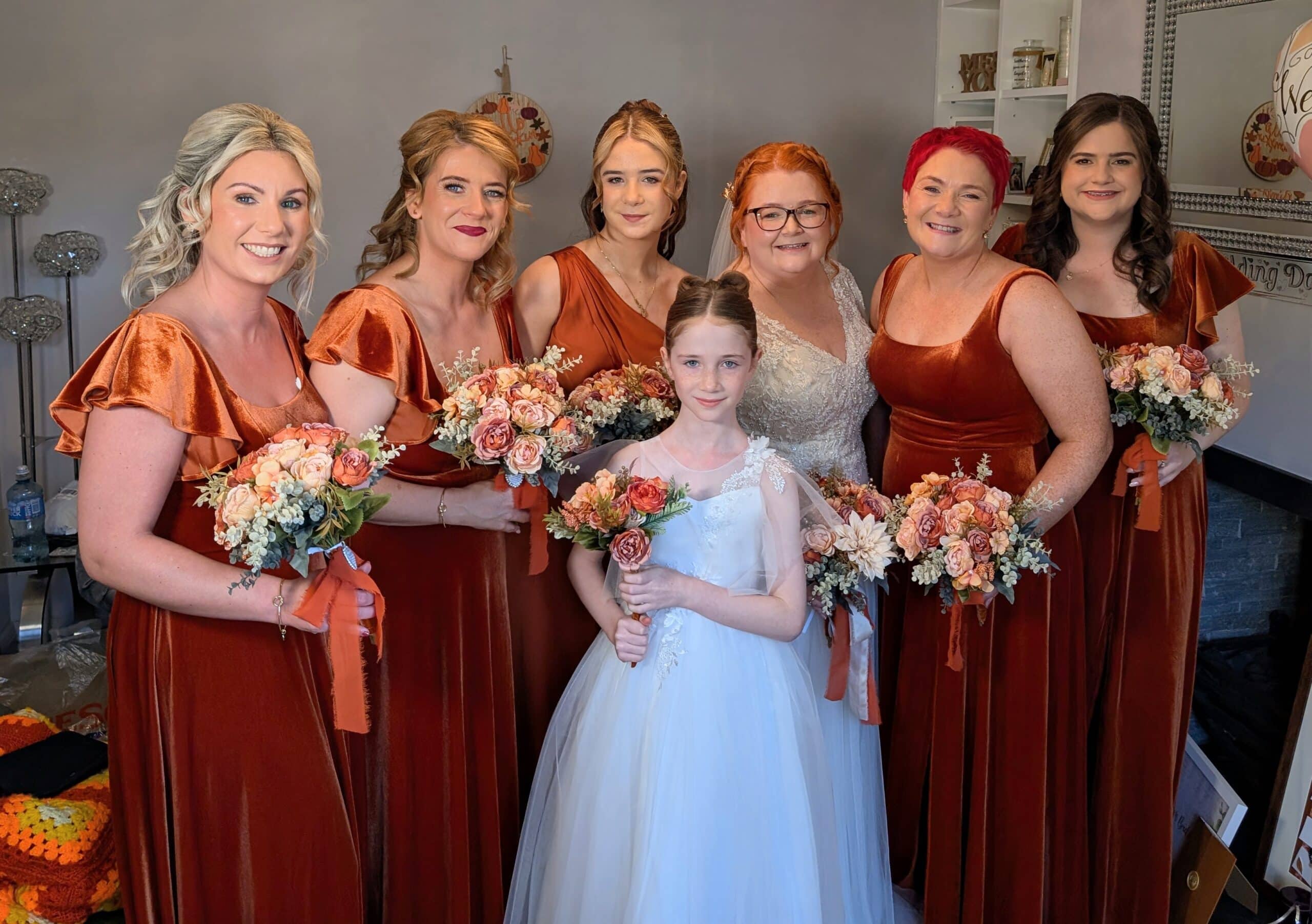 Bridal party with bridesmaids in matching copper dresses and young girl holding bouquets, smiling indoors.