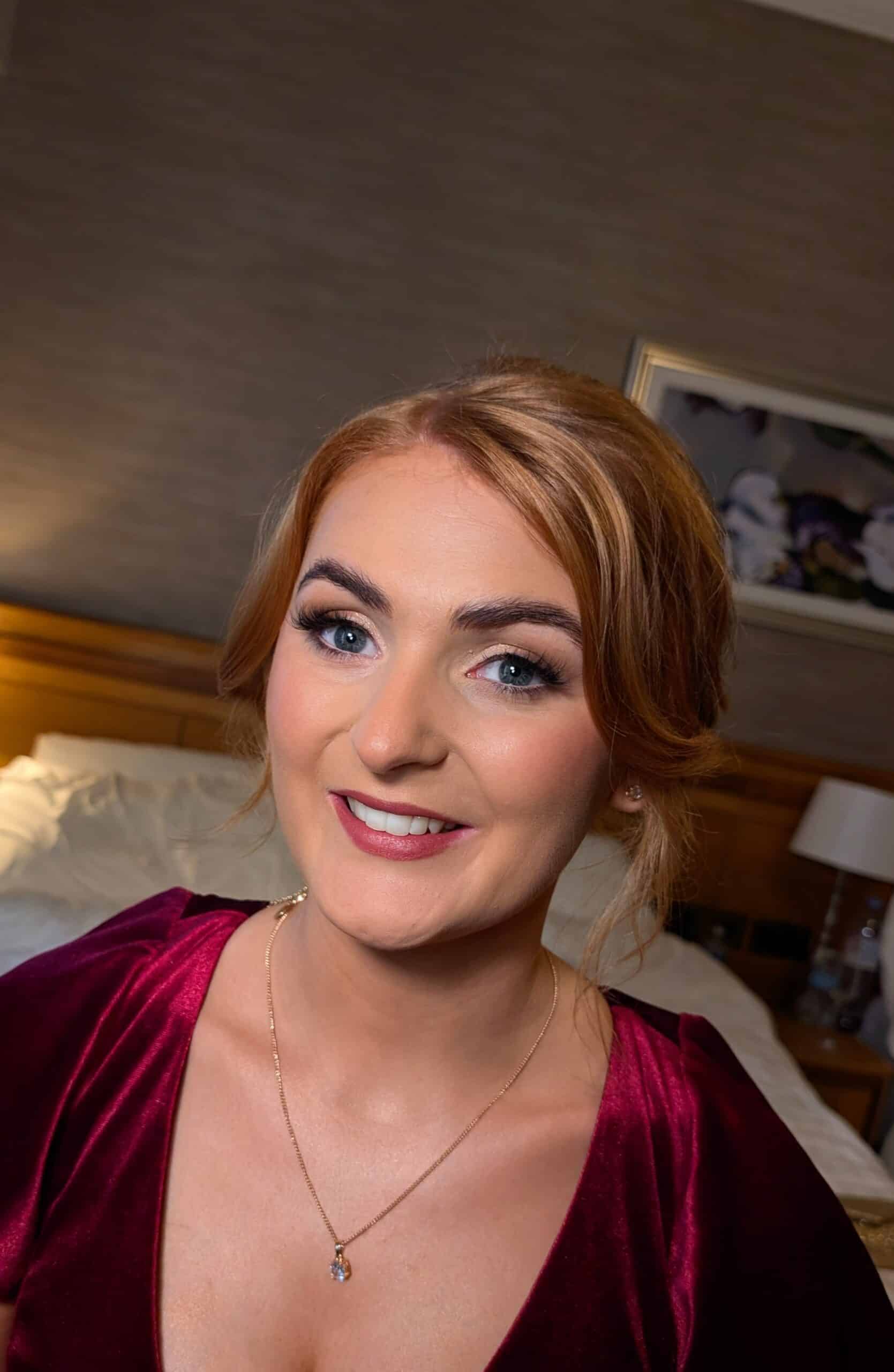 Smiling woman with styled hair and burgundy dress, necklace visible, in a warmly lit room.