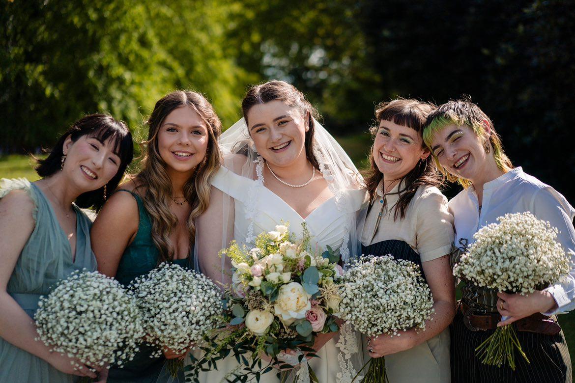 Bride and bridesmaids smiling, holding bouquets of flowers outdoors.