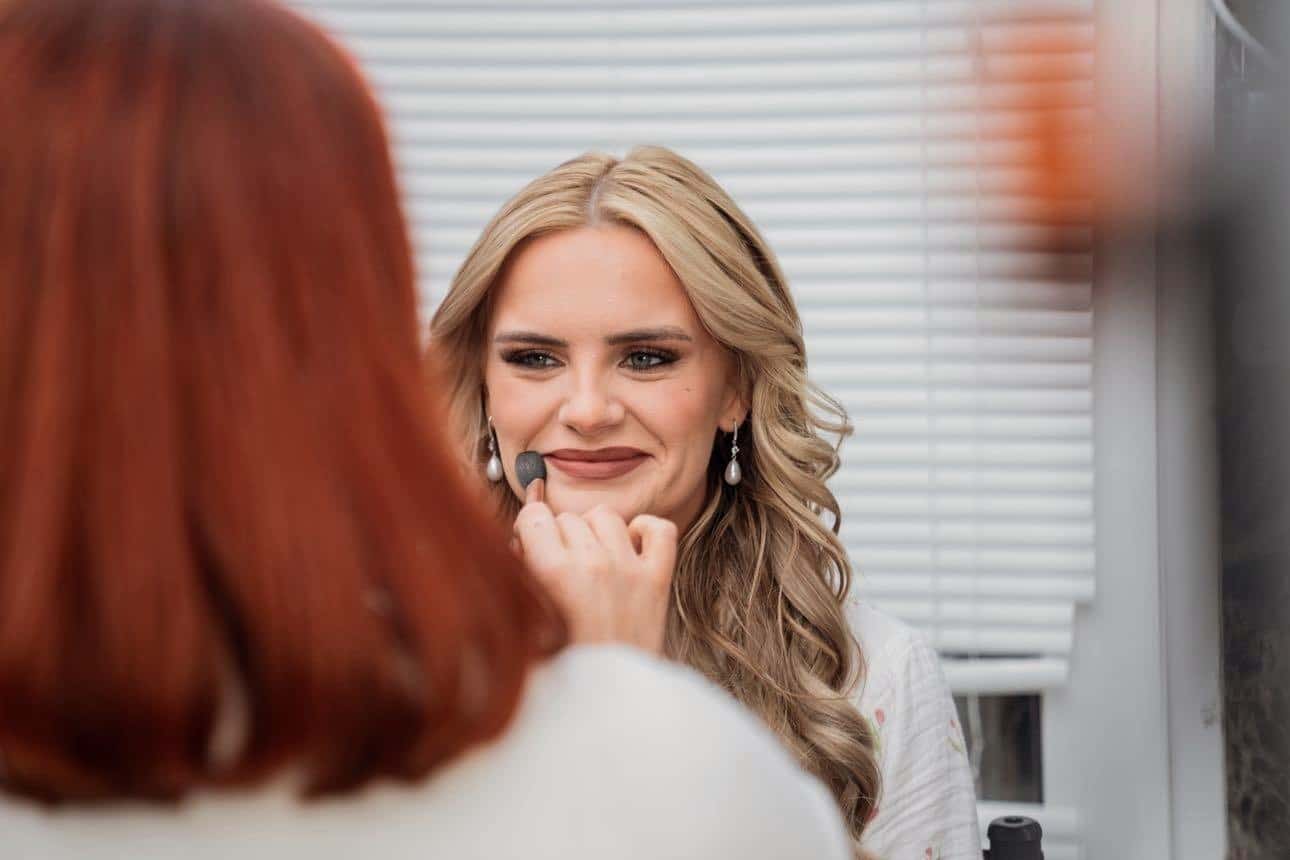 Woman having makeup applied, smiling at the makeup artist in front of a window with blinds.