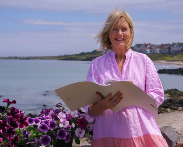 Woman holding a folder, standing near coastal water with flowers nearby.