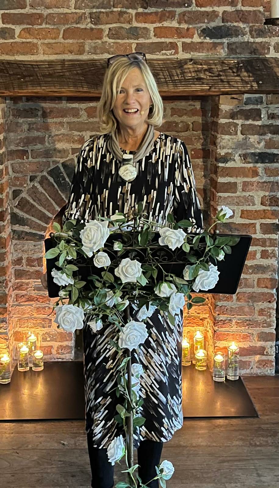 Woman standing at a decorated podium with white flowers in a brick-walled room.