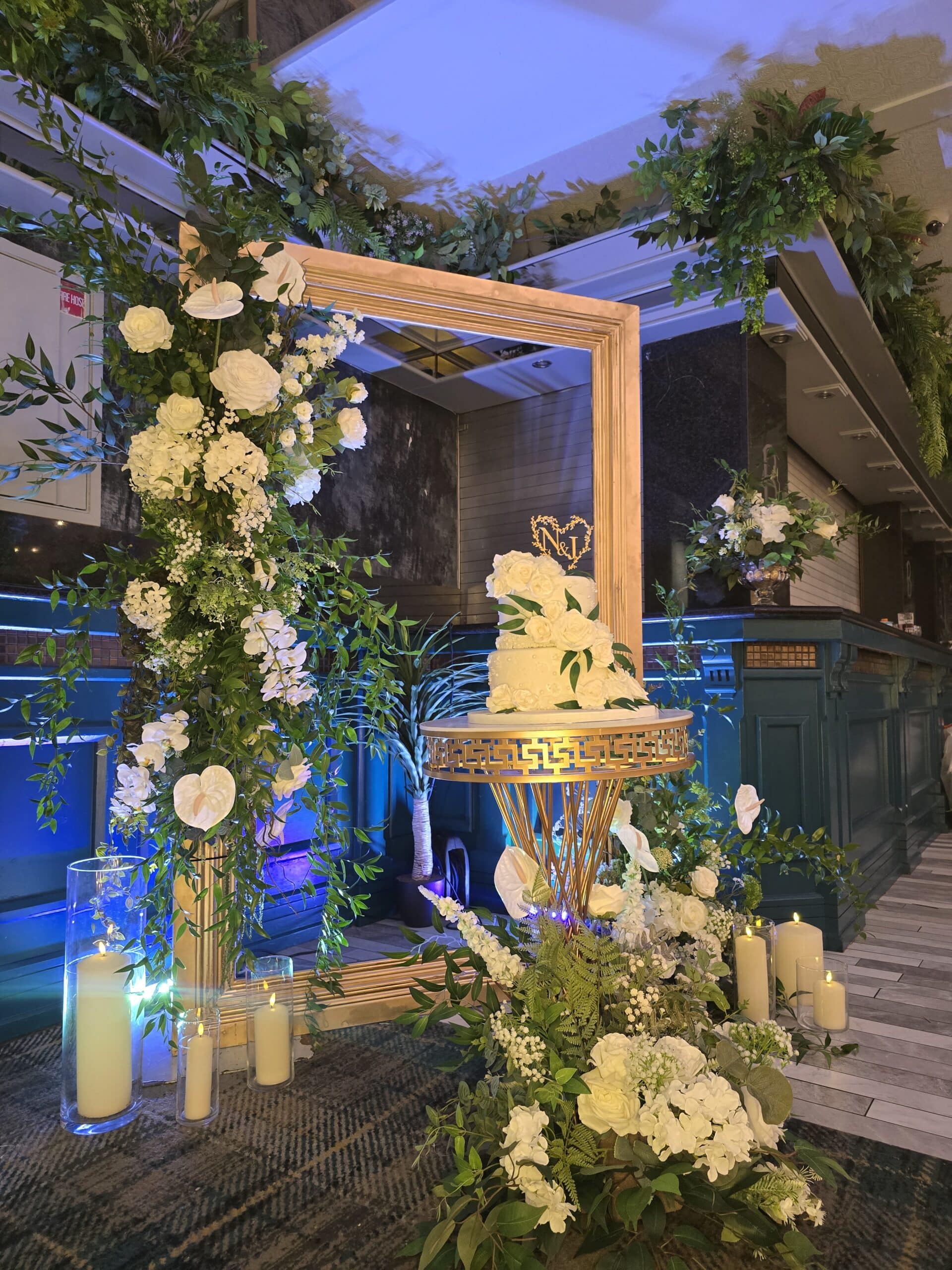 Elegant wedding cake display with floral arrangements, candles, and a decorative frame in a softly lit venue.