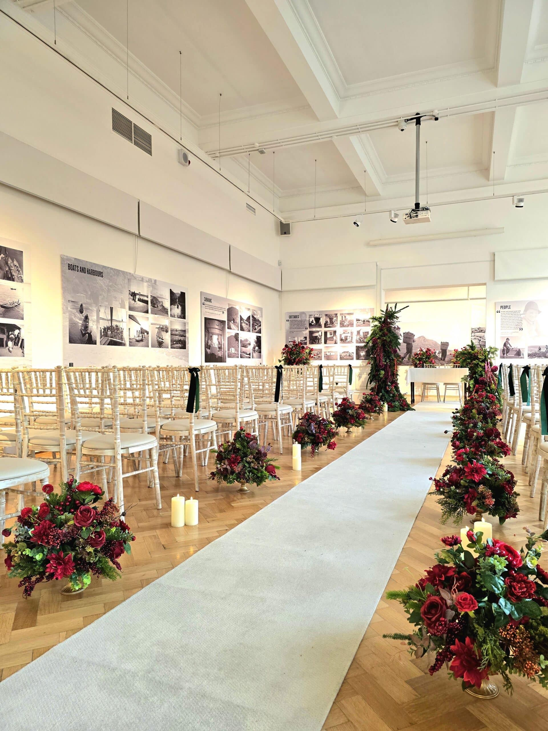 Elegant wedding aisle with white chairs, floral arrangements, and candles in a spacious gallery setting.