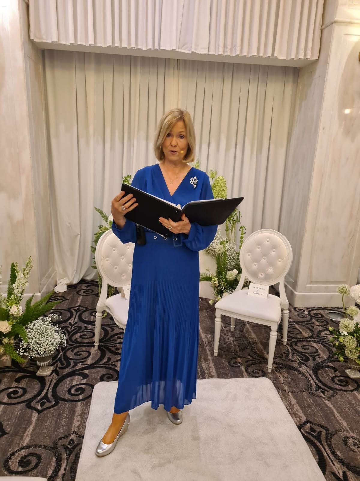 Woman in blue dress reading from a book in an elegant room with floral decorations and white chairs.