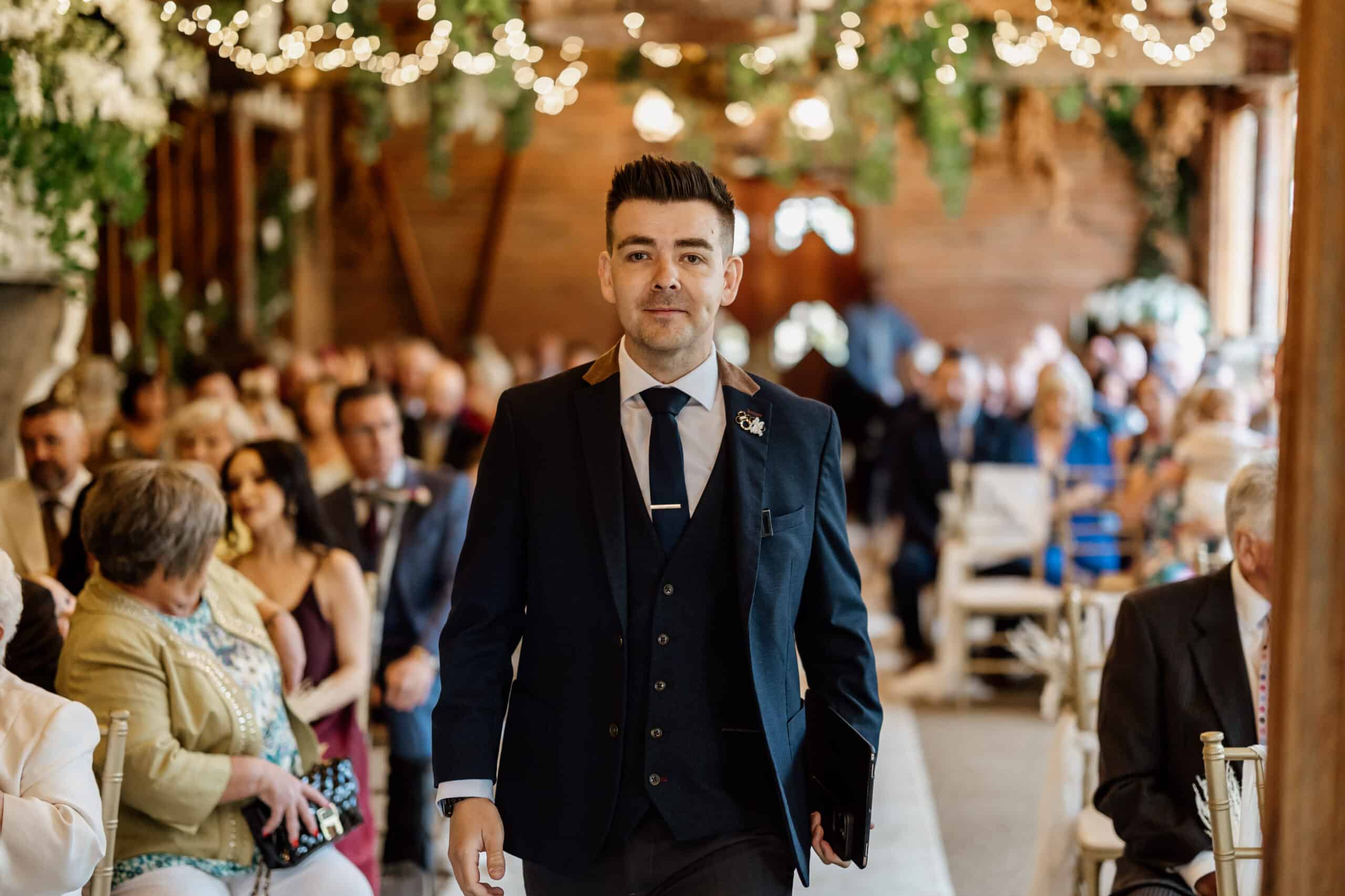 Man in formal attire walking down an aisle at an indoor event with guests seated.