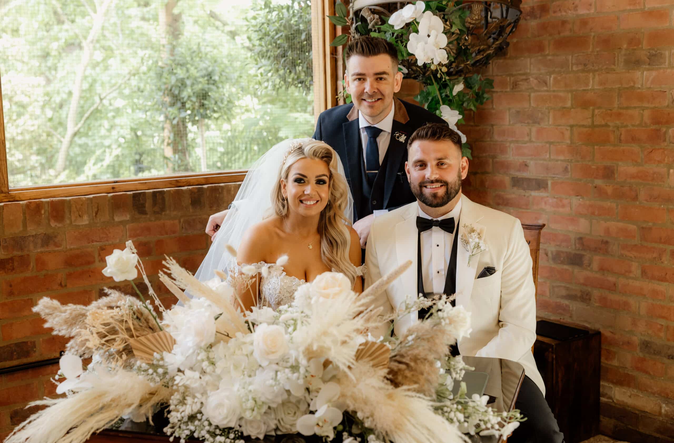 Bride and groom smiling with a friend, surrounded by flowers indoors.