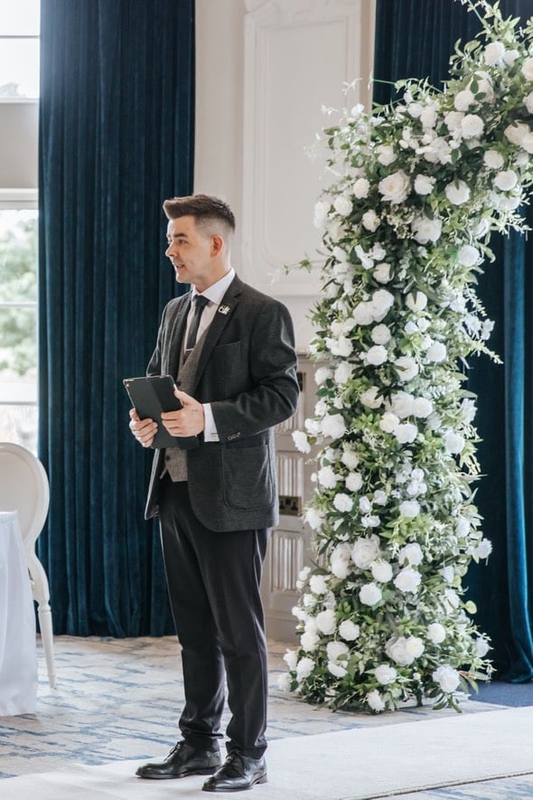 Man in formal attire holding a tablet, standing near a floral arch in an elegant room.