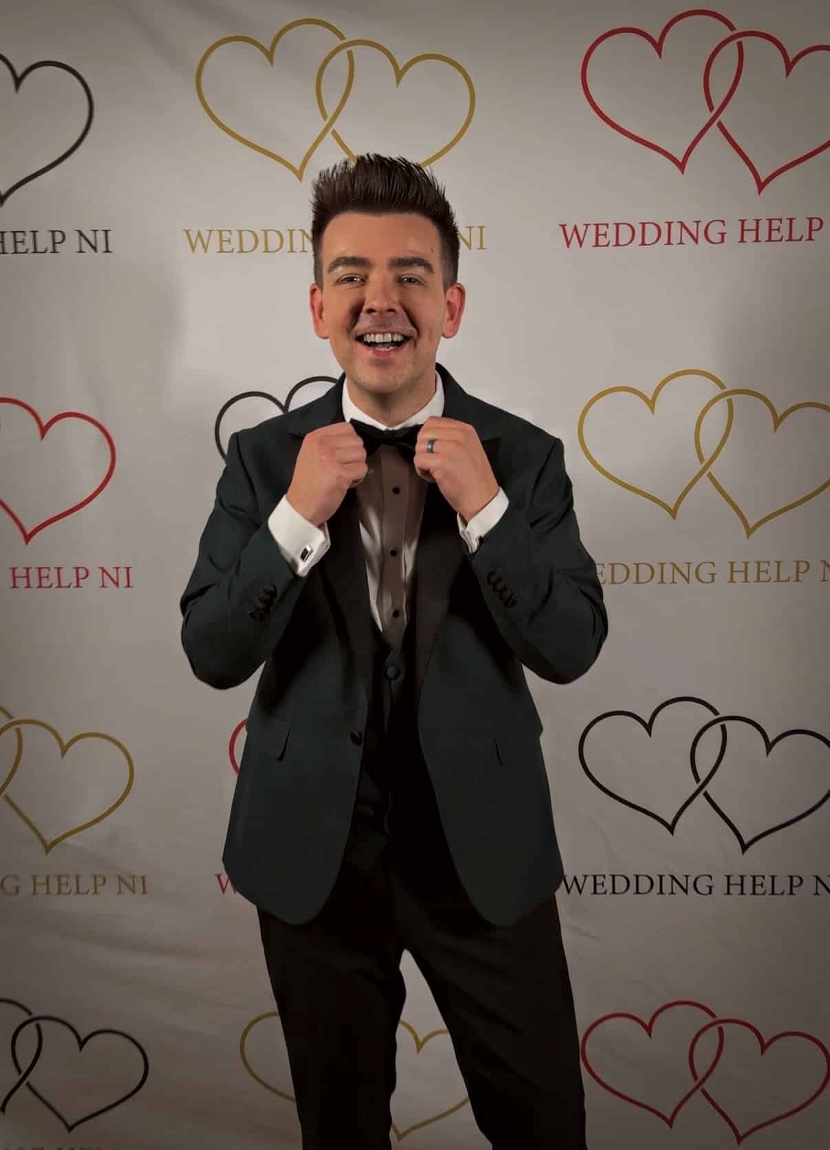 Man in a suit adjusting his bow tie, smiling in front of a "Wedding Help NI" backdrop with heart designs.